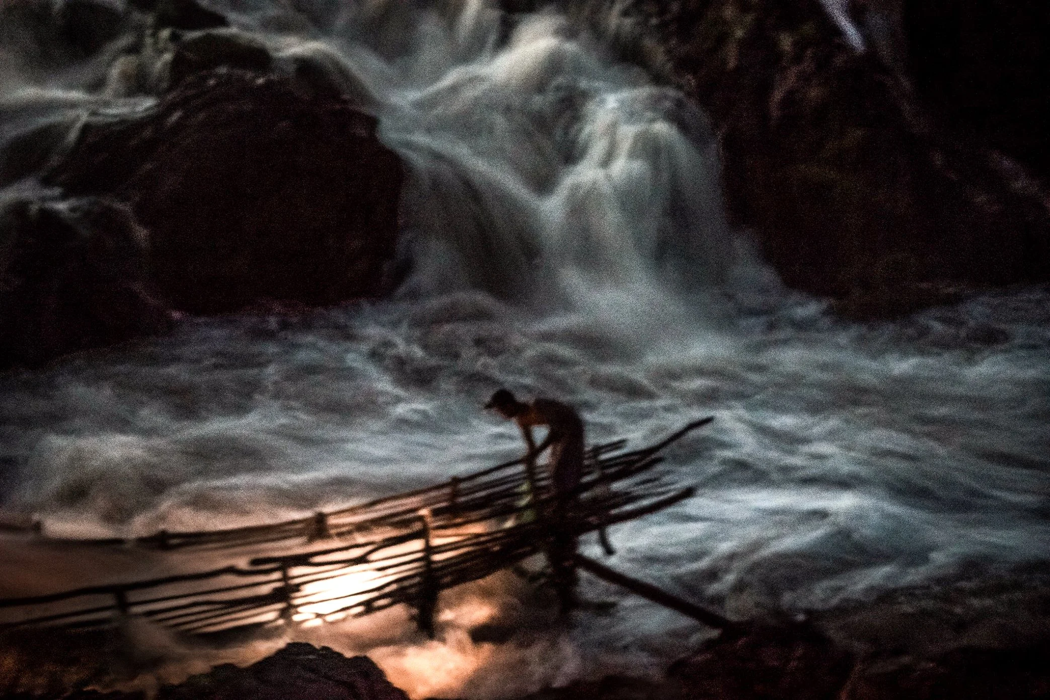 A fisherman checks his Li trap at night in Si Phan Don, silhouetted against the white force of the Mekong. During the migration season, the work continues after dark, when fish may enter the trap with the changing current and the structure must be cl