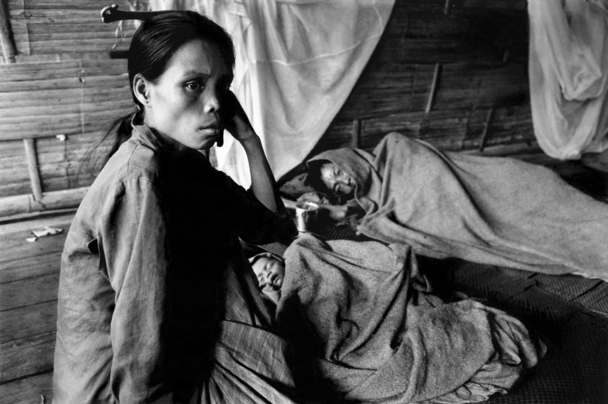 Black and white photograph of a woman sitting beside a bed with two sick or injured people lying under blankets in a rustic wooden room.