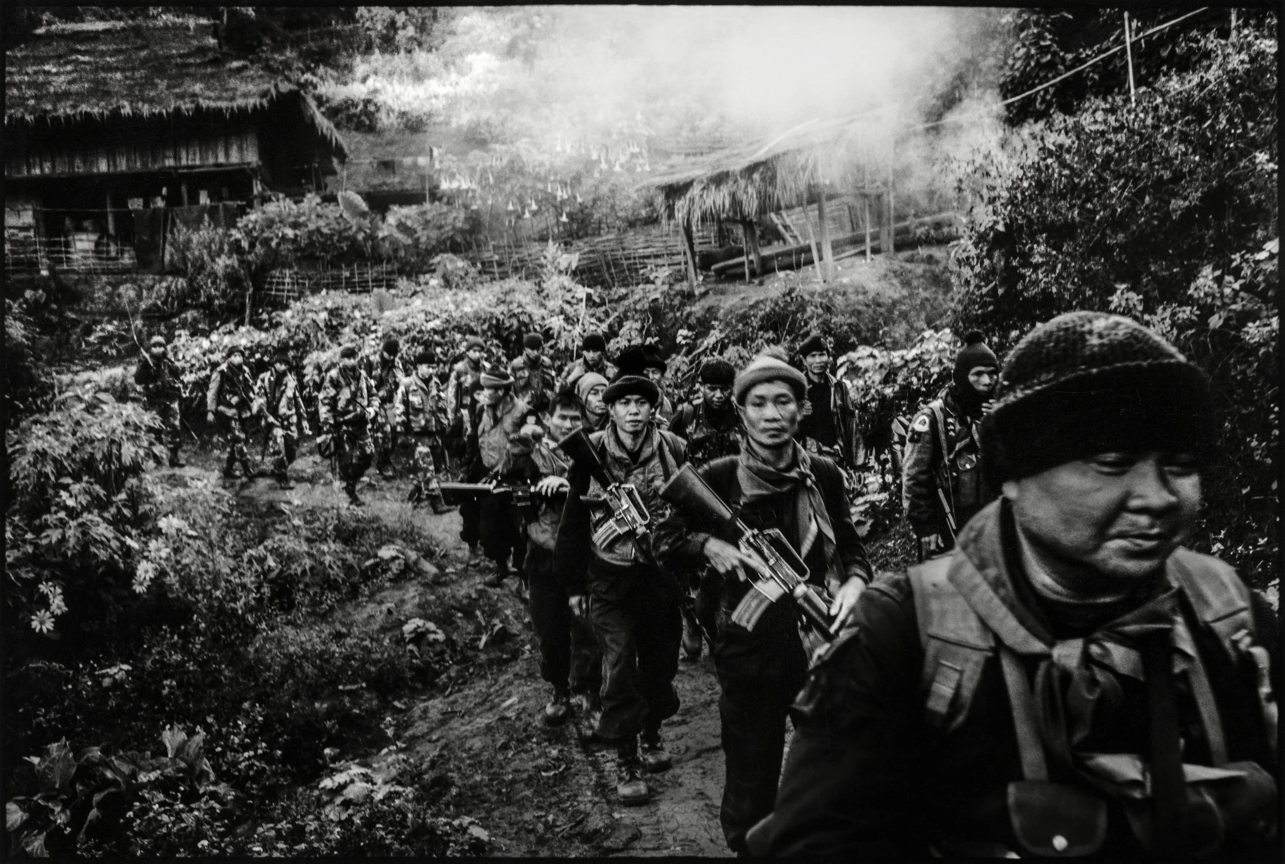 A group of soldiers carrying rifles walking along a dirt path through a rural, mountainous landscape with traditional huts and lush vegetation.