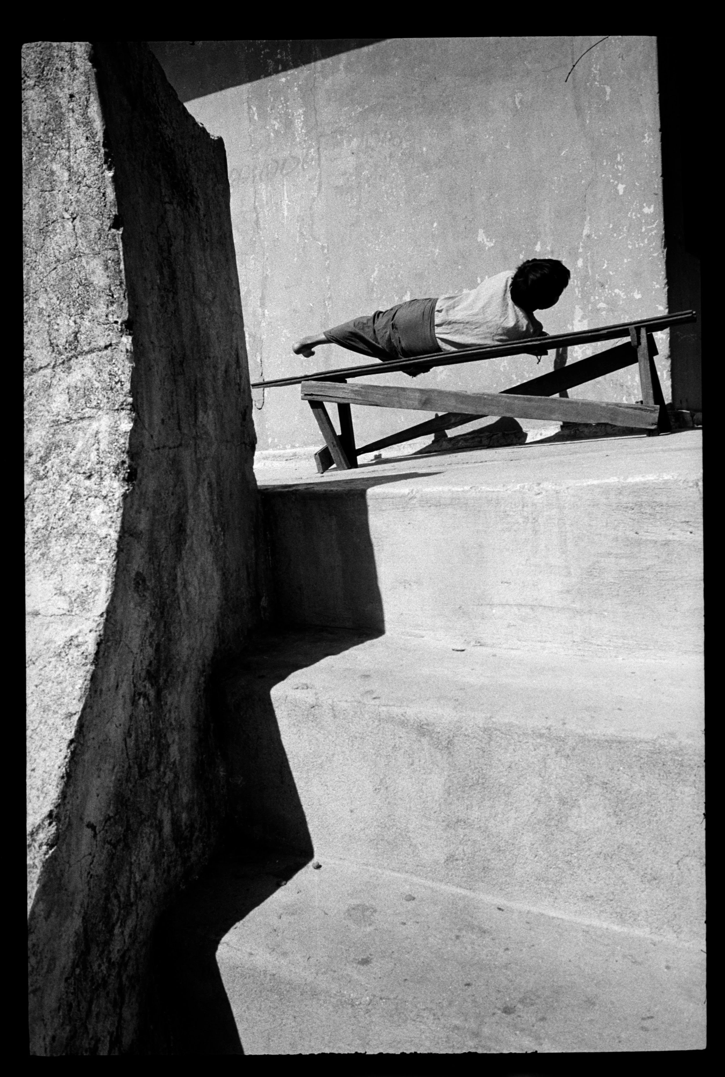 A black and white photo of a man lying sideways on a wooden bench, with his head against a wall and his legs extended. The bench is tilted at an angle, and the photo is taken from a low perspective, showing the shadow of a rock formation on the ground.