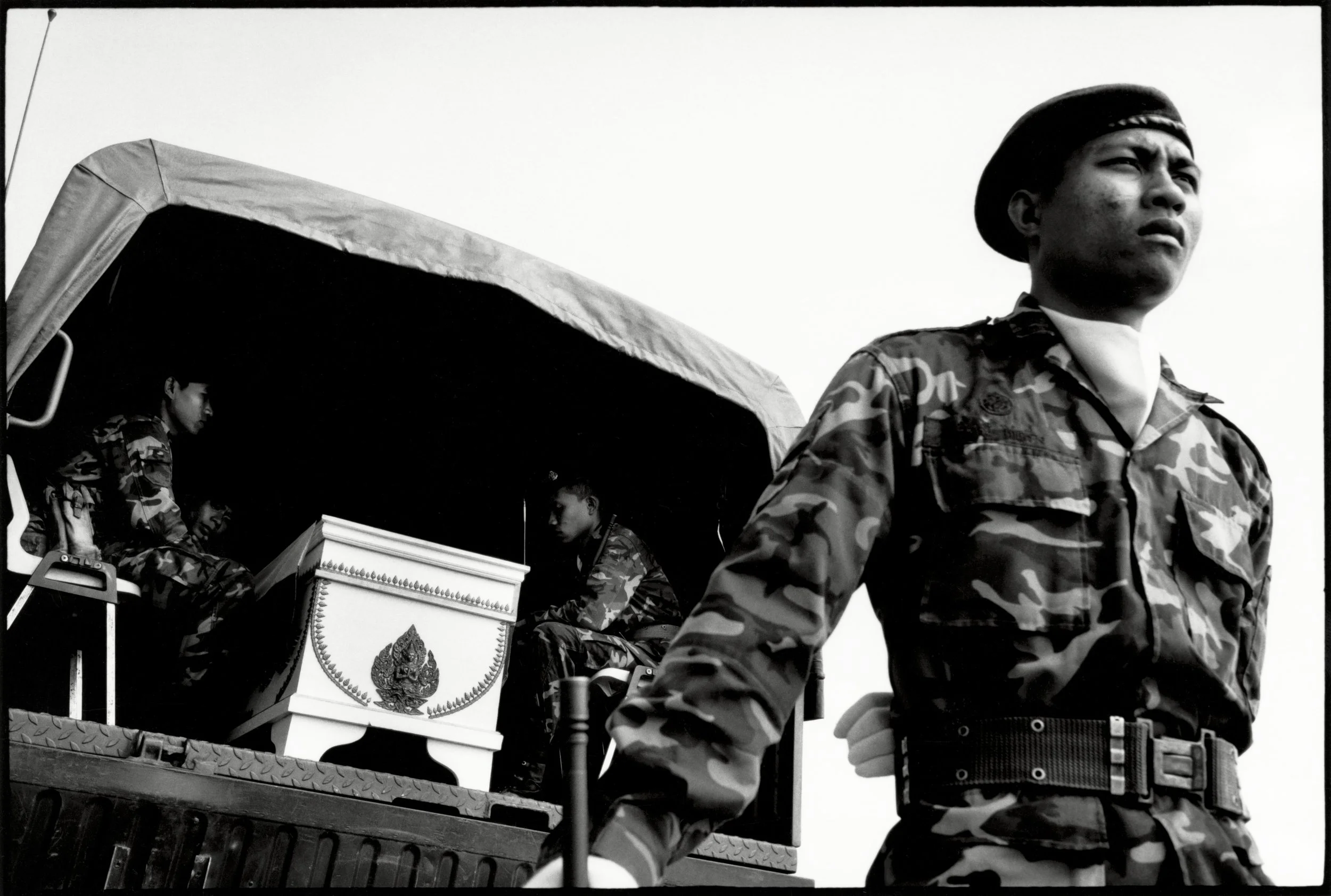 A soldier in camouflage uniform standing in front of a military vehicle with other soldiers seated inside, partially visible, in the background.