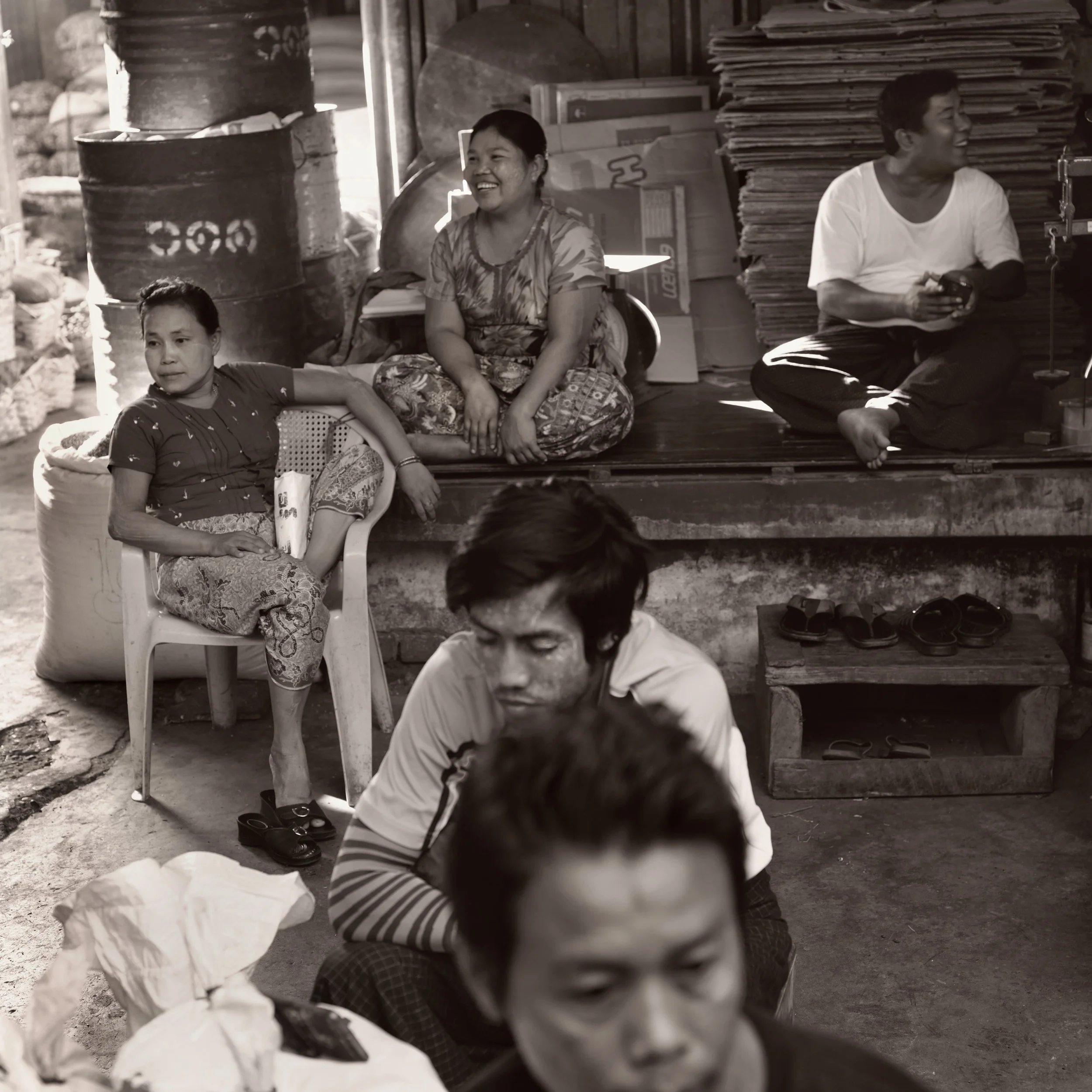 A group of people sitting and standing in a rustic setting with stacked cardboard boxes and barrels in the background, some smiling and engaging with others.