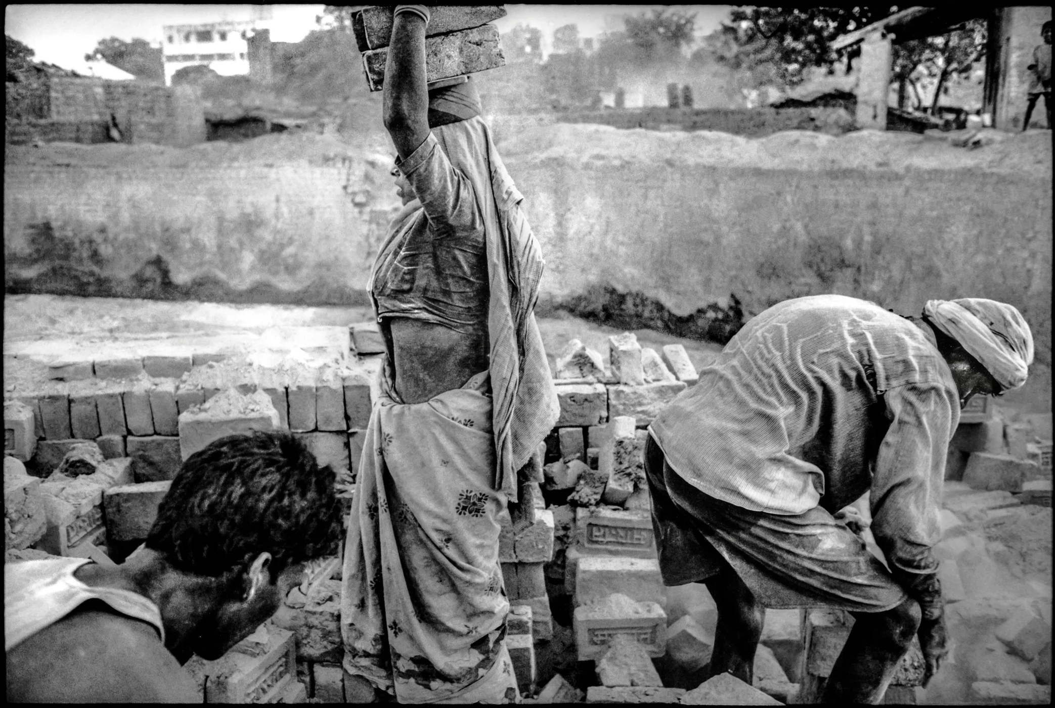 Patna, Bihar, India - April 2003.
Under the harsh April sun, workers move bricks inside a kiln pit where heat rises from the ground and dust hangs in the air. Each load is carried by hand, one stack after another, in a cycle of labor shaped by povert