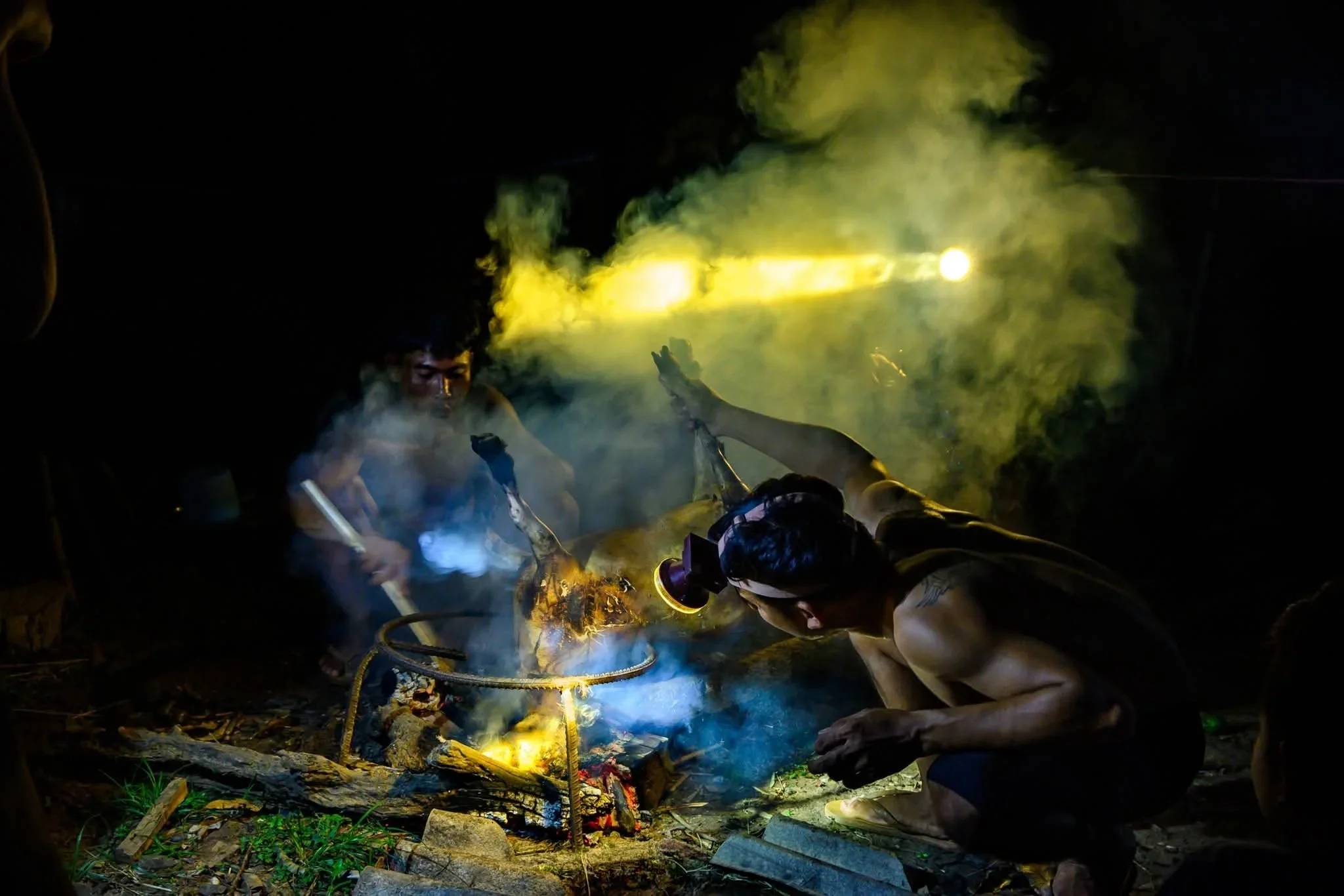 Two individuals are crouching near a small outdoor fire with yellow-green smoke, working together to tend to or build the fire, surrounded by stones and wood pieces.