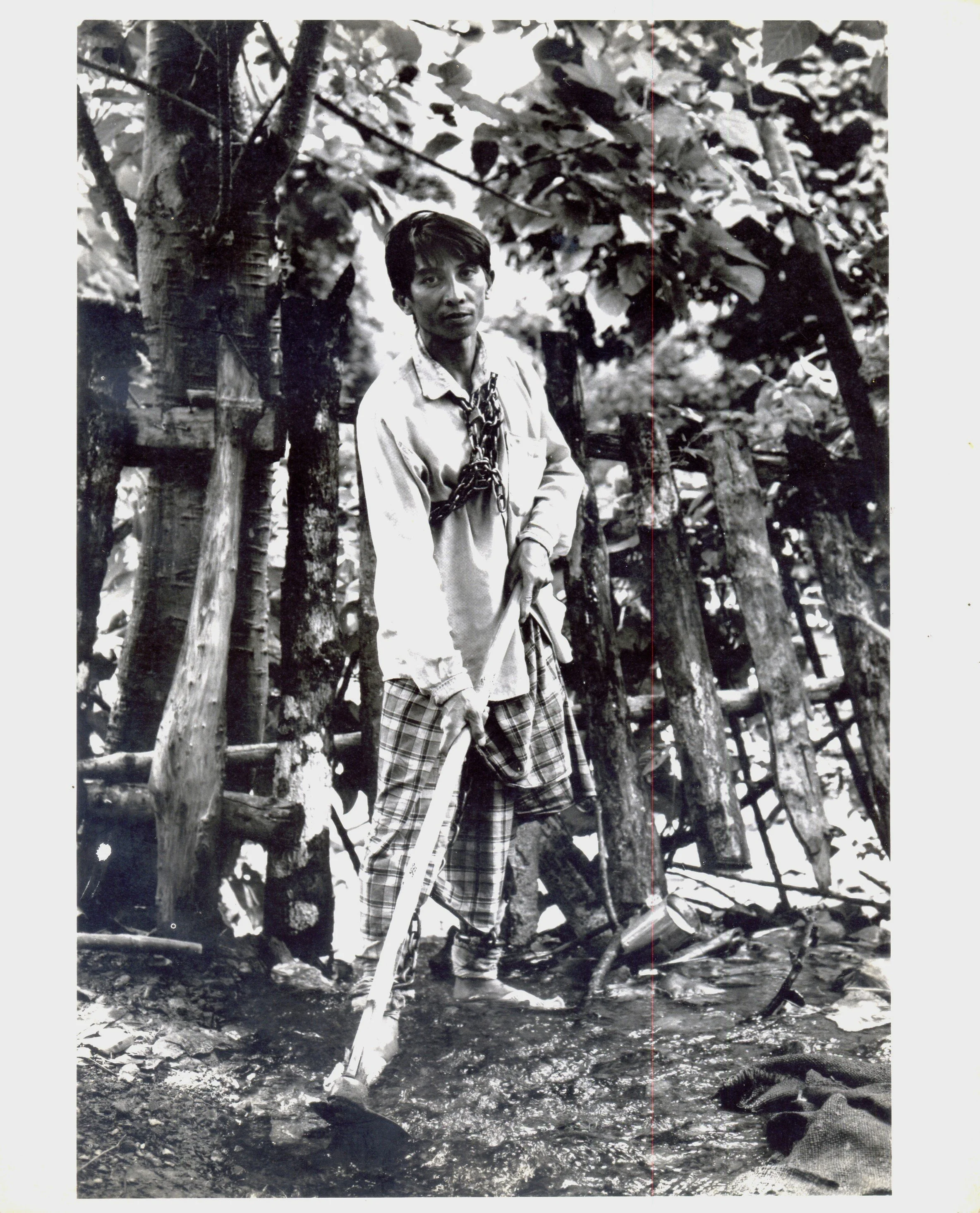 A man standing in a stream with a stick, wearing a long-sleeved shirt and plaid pants, with a chain around his neck, in front of a wooden fence and trees.