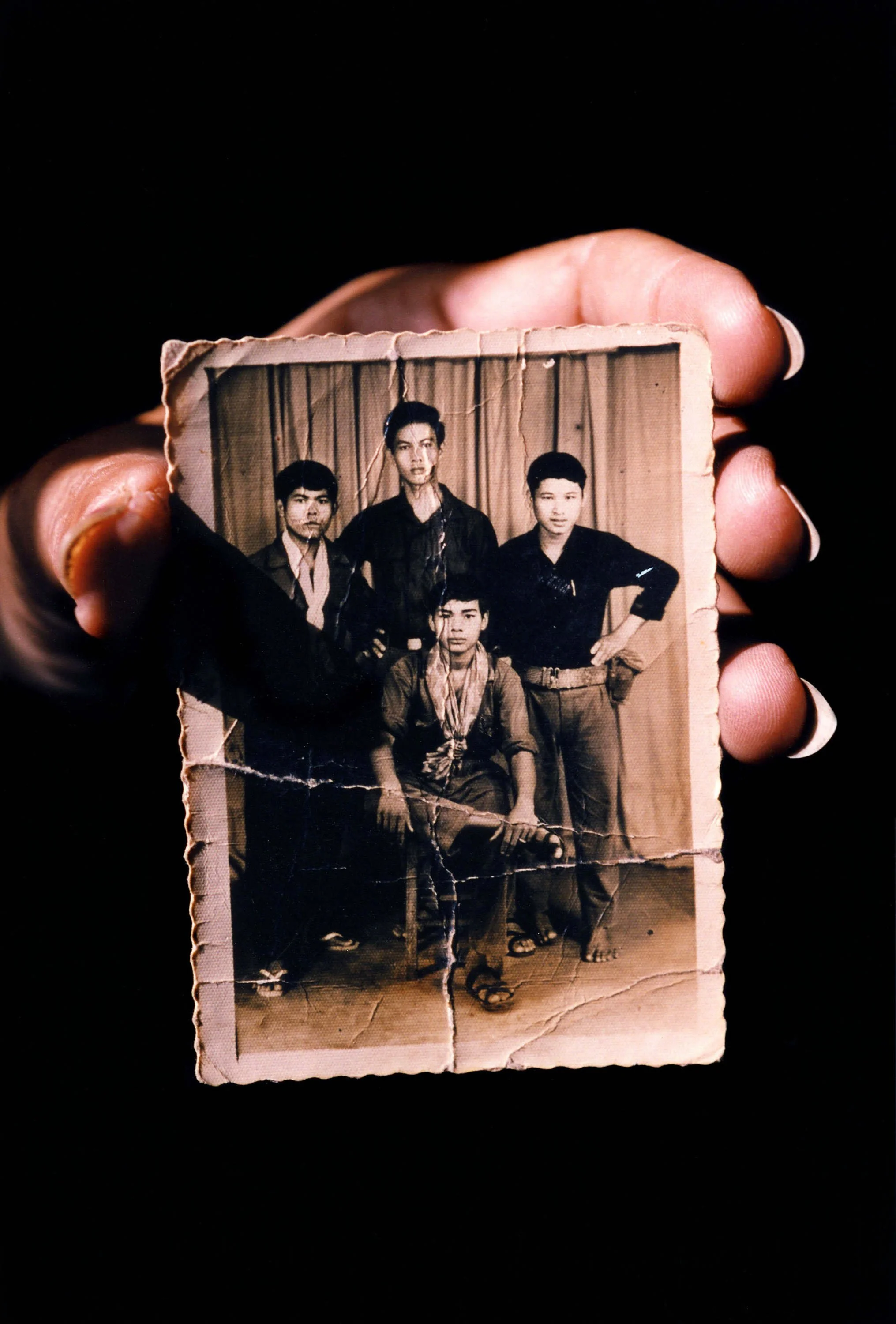 A worn, black-and-white photograph of five young men posing against a curtain backdrop, held by a person with dark skin and manicured nails.