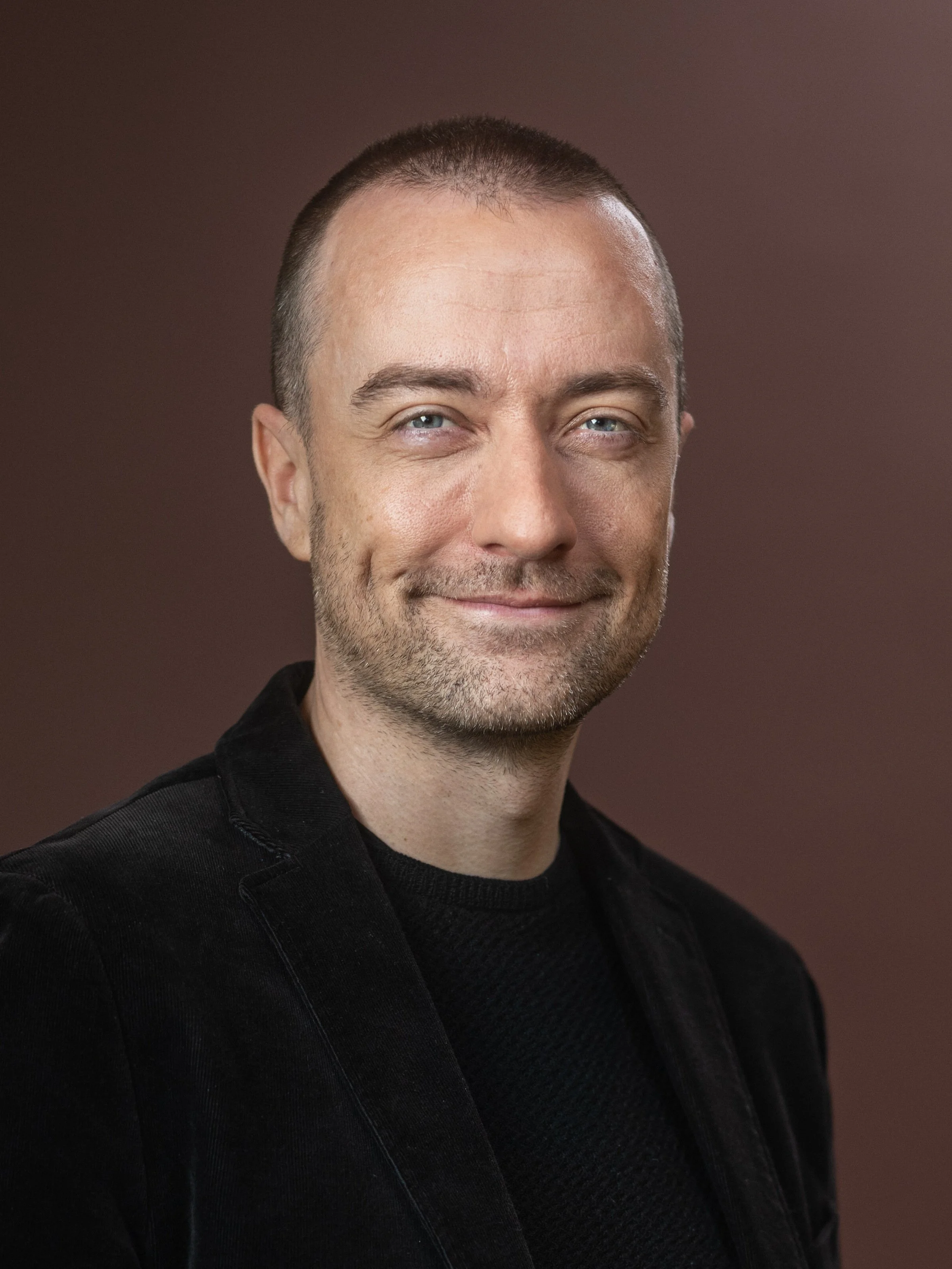 A man with short brown hair and light skin, wearing a black blazer and shirt, smiling softly against a brown background.