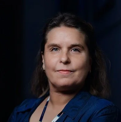 A woman with dark hair and a slight smile, wearing a dark blue shirt, against a dark background.