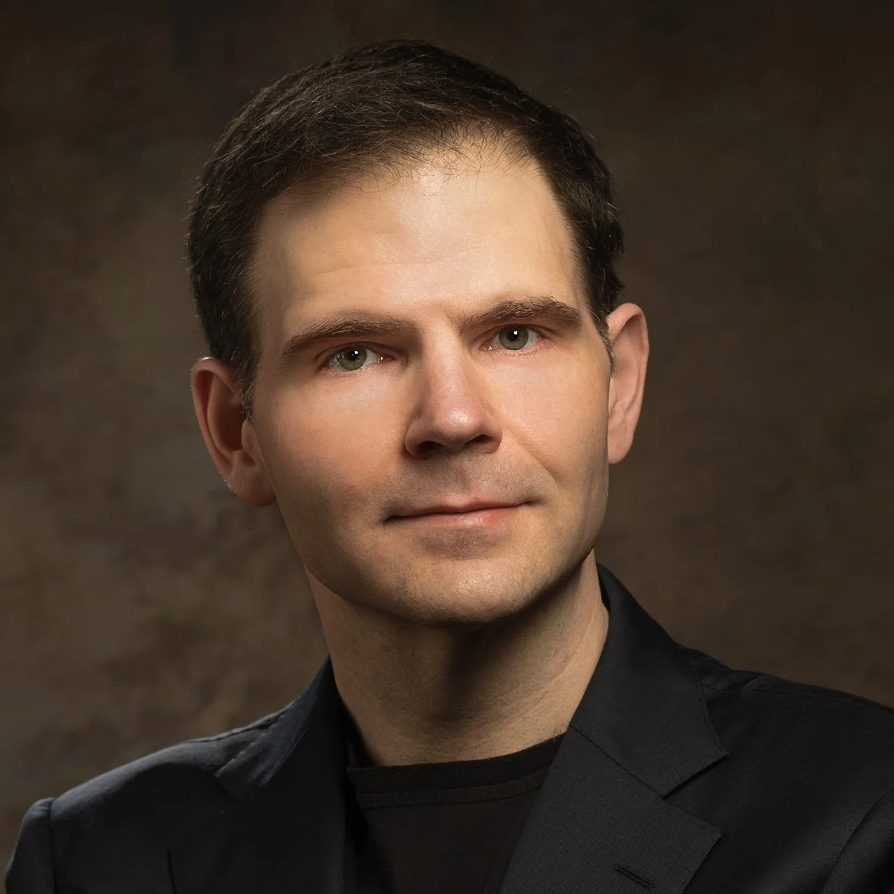 Portrait of a man with short dark hair, light skin, and green eyes, wearing a black blazer against a dark brown background.