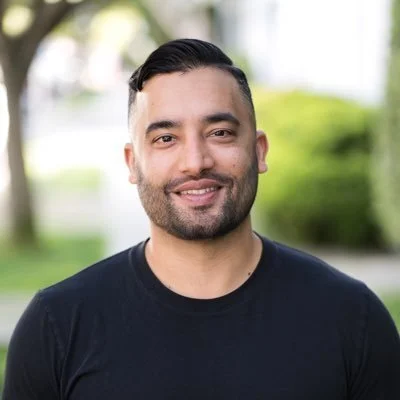 A man with dark hair and a beard smiling outdoors with greenery in the background.