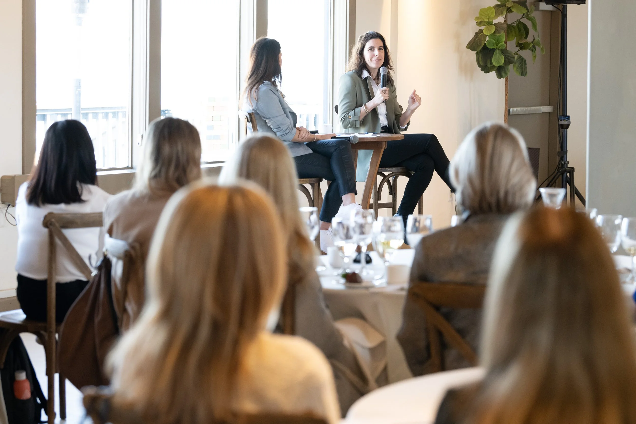 A woman speaking into a microphone during a panel discussion at a conference, with an audience seated at tables, in a bright room with large windows and a potted plant in the background.