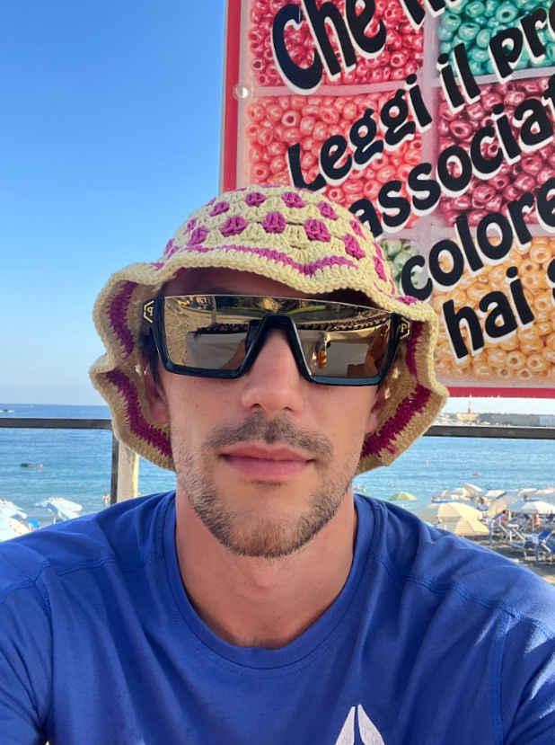 Uomo con cappello di paglia e occhiali da sole seduto vicino al mare con ombrelloni e sedie in spiaggia, sullo sfondo un cartellone colorato.
