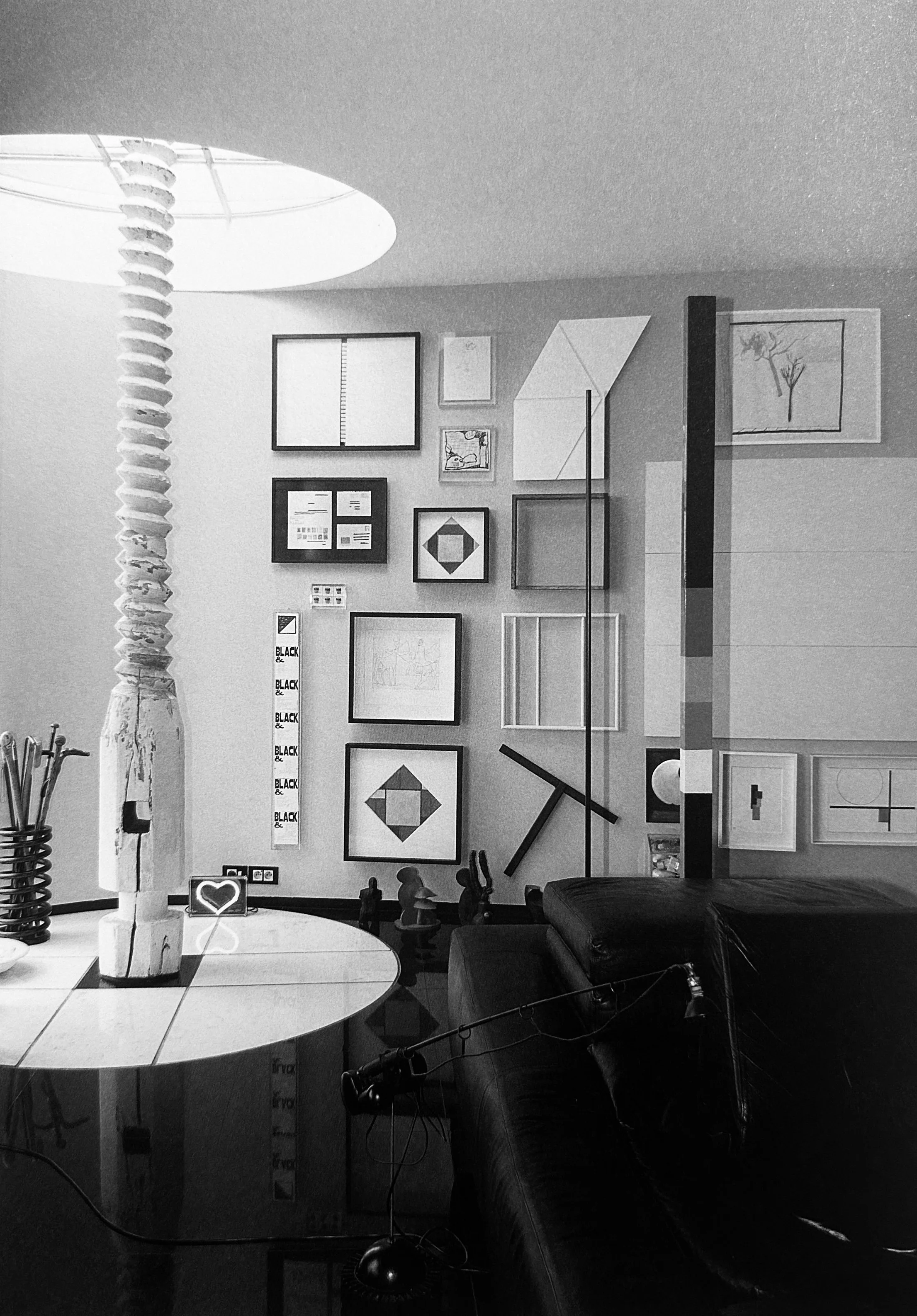 Een zwart-wit foto van een woonkamer met een muur vol verschillende kunstwerken en lijsten, een tafel met een sculptuur en een bank met een lamp en decoraties.