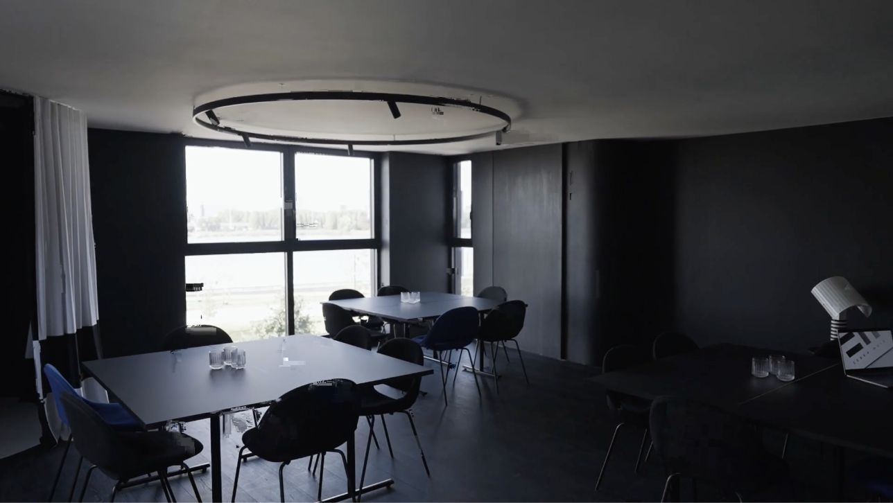 De  vergaderruimte in het Huis van Roosmalen met grote ramen, een ronde verlichtingsring uit het plafond, en meerdere tafels met stoelen, enkele met waterglazen erop.