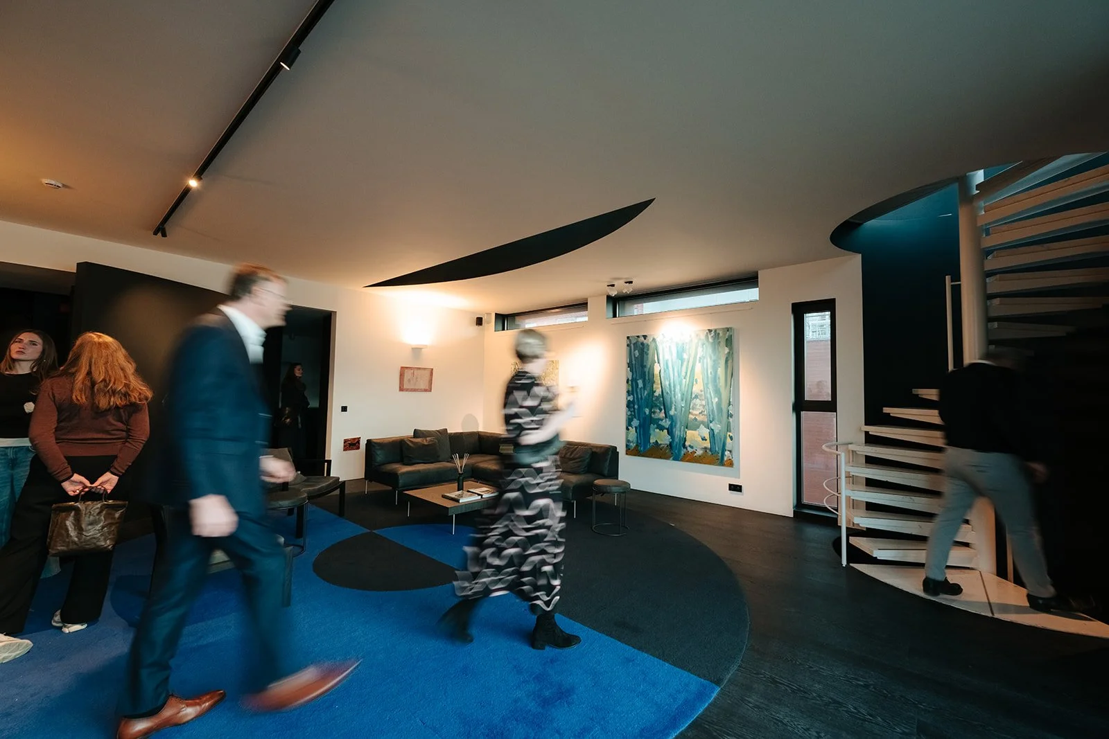 Binnenruimte van het Huis van Roosmalen met mensen, een zwarte bank, een ronde Yves Klein-blauwe en zwarte tapijt, een trap naar rechts, en kunst aan de muur, verlichte muren en ramen.