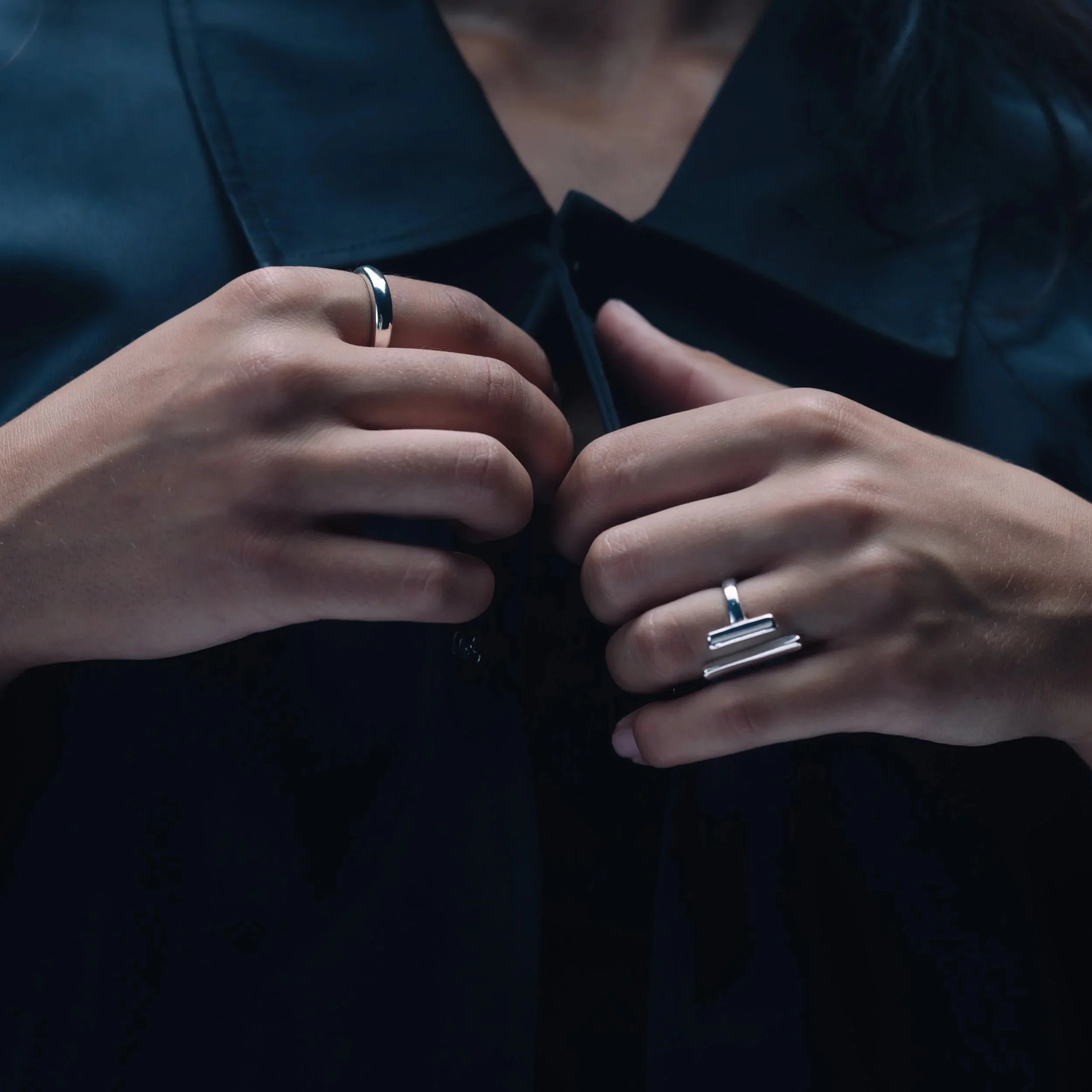 Nahaufnahme von Händen mit silbernen Ringen, die eine schwarze Jacke anknöpfen.