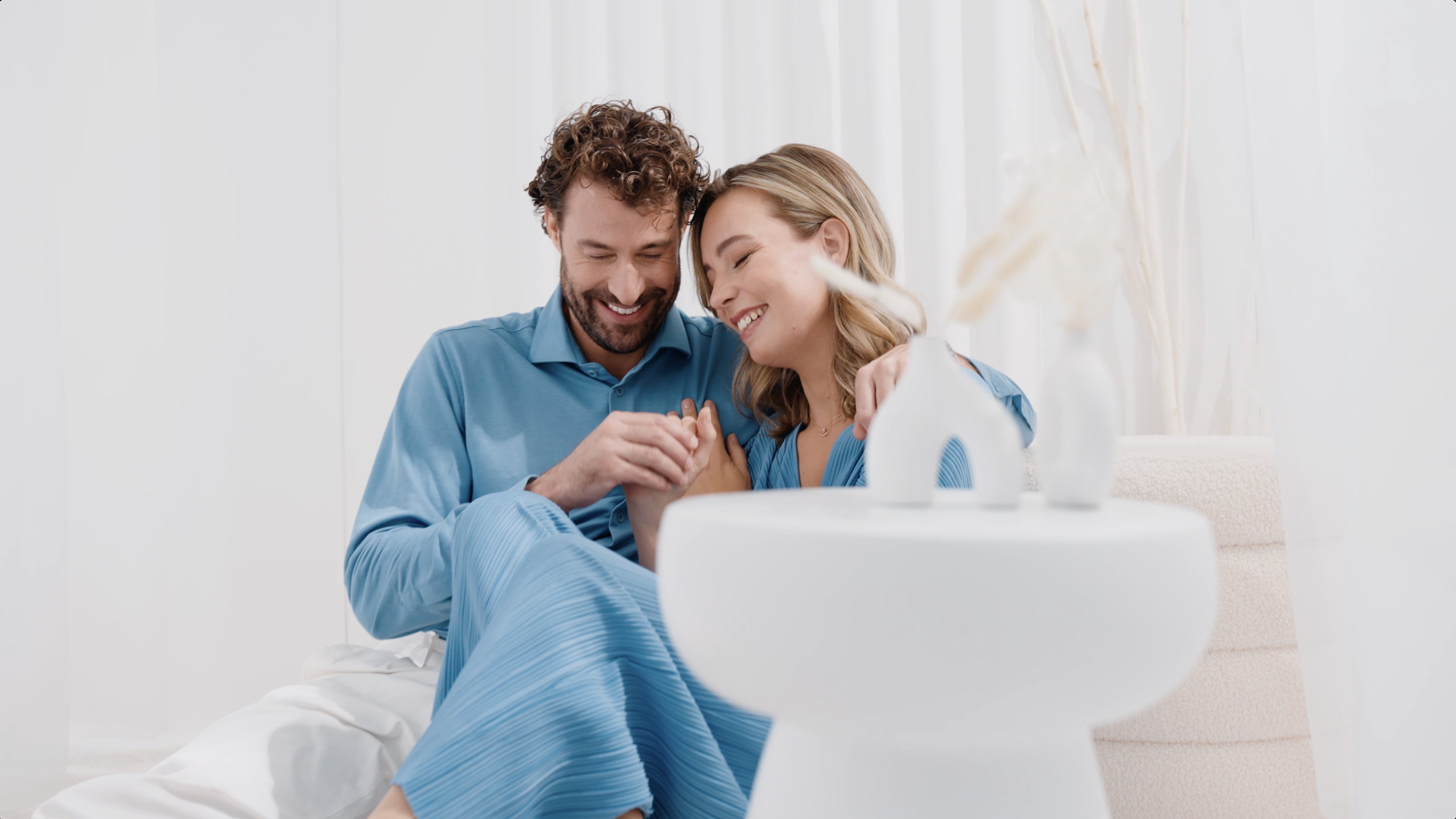 Ein lachendes Paar sitzt auf einem Bett, das in einem hellen Raum mit weißen Wänden sitzt, und hält sich an den Händen, während sie sich anlächeln. Im Vordergrund ist eine weiße Vase mit trockenen Pflanzen auf einer Ablage.