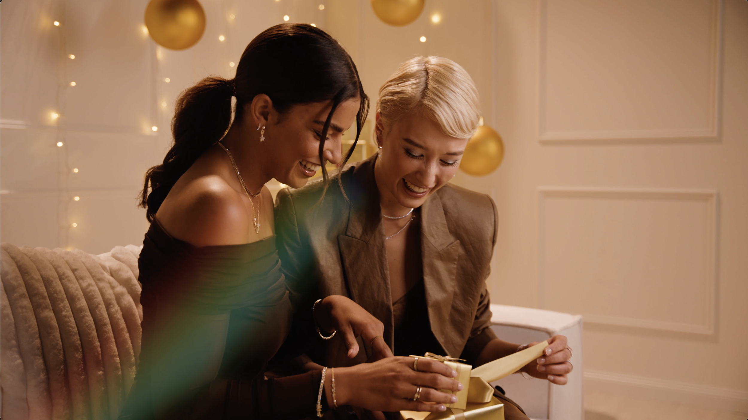 Zwei Frauen lachen und öffnen gemeinsam ein Geschenk, umgeben von festlicher Dekoration mit goldenen Kugeln und Lichterketten.