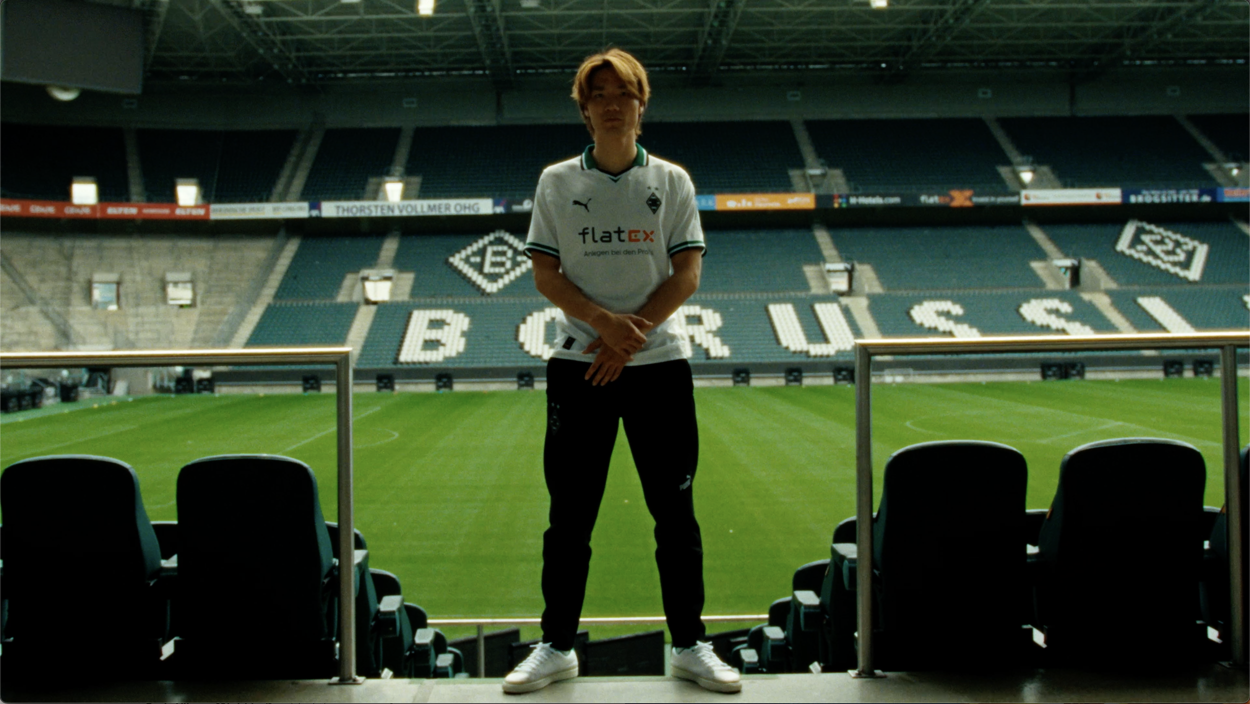 Person steht in einem leeren Stadion auf der Tribüne, trägt ein weißes Fußballtrikot und schwarze Hosen, im Hintergrund sind leere Sitzreihen und ein grünes Spielfeld sichtbar.