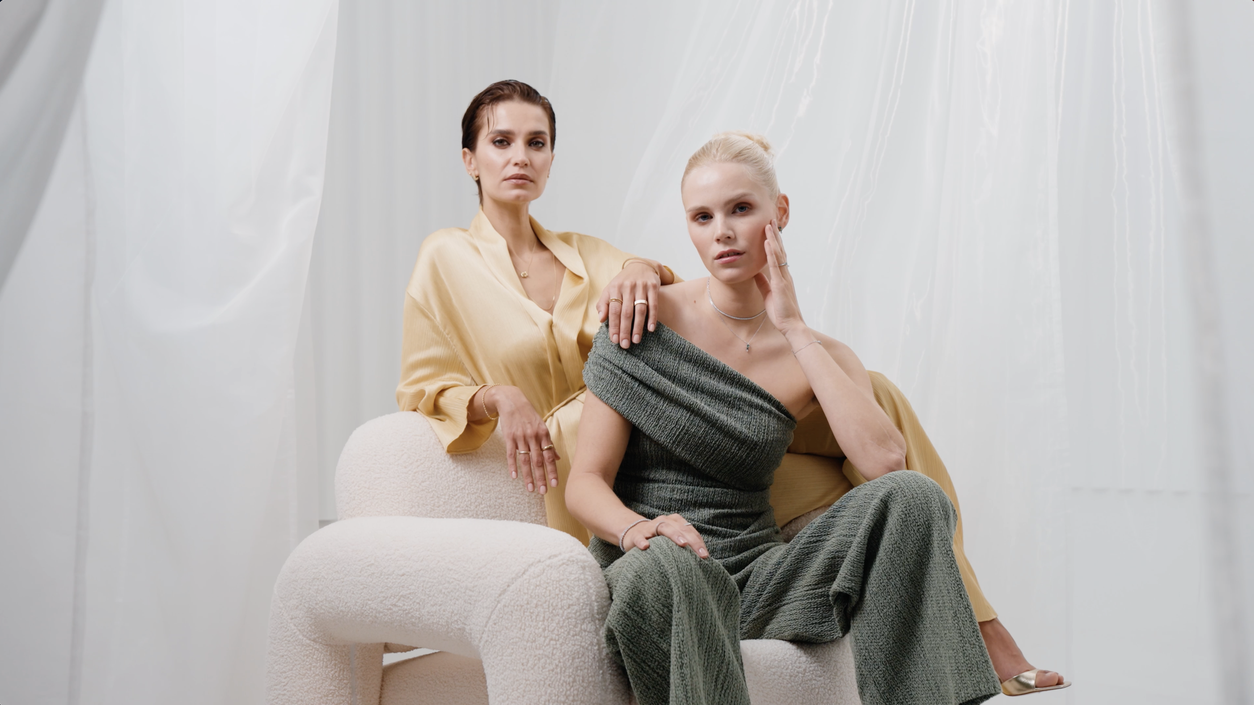 Zwei Frauen sitzen auf einem weißen Sitz, umgeben von weißen Vorhängen, in eleganten Outfits und mit neutralem Gesichtsausdruck.