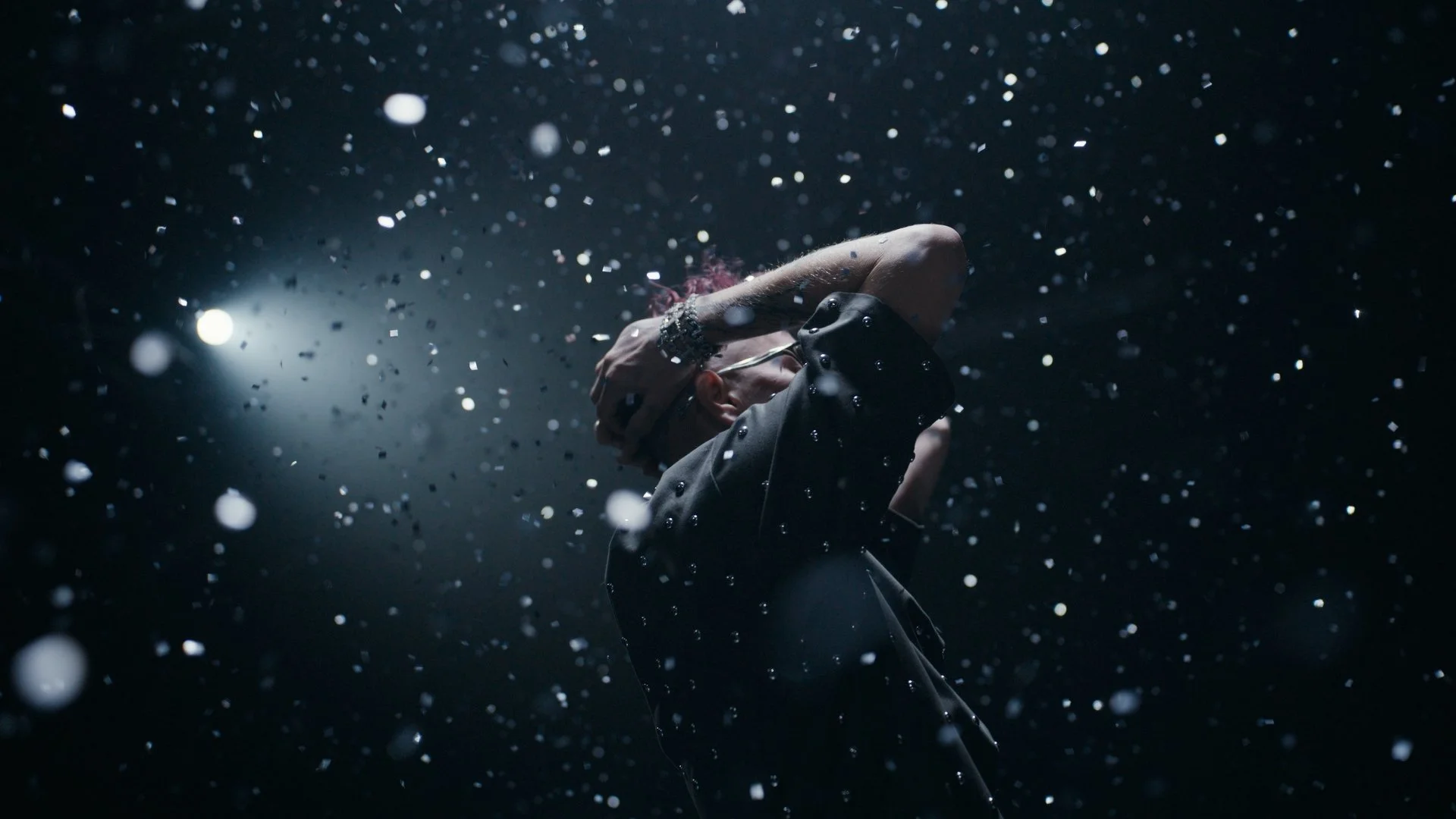 Eine Person steht im Dunkeln, umgeben von fallendem Schnee oder Konfetti, mit einer Hand am Kopf, bei Nacht, mit einer hellen Lichtquelle im Hintergrund.