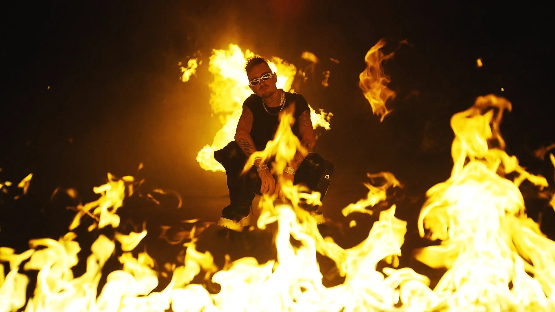 Mann mit Tattoos, Sonnenbrille und Schmuck steht vor Flammen bei Nacht.