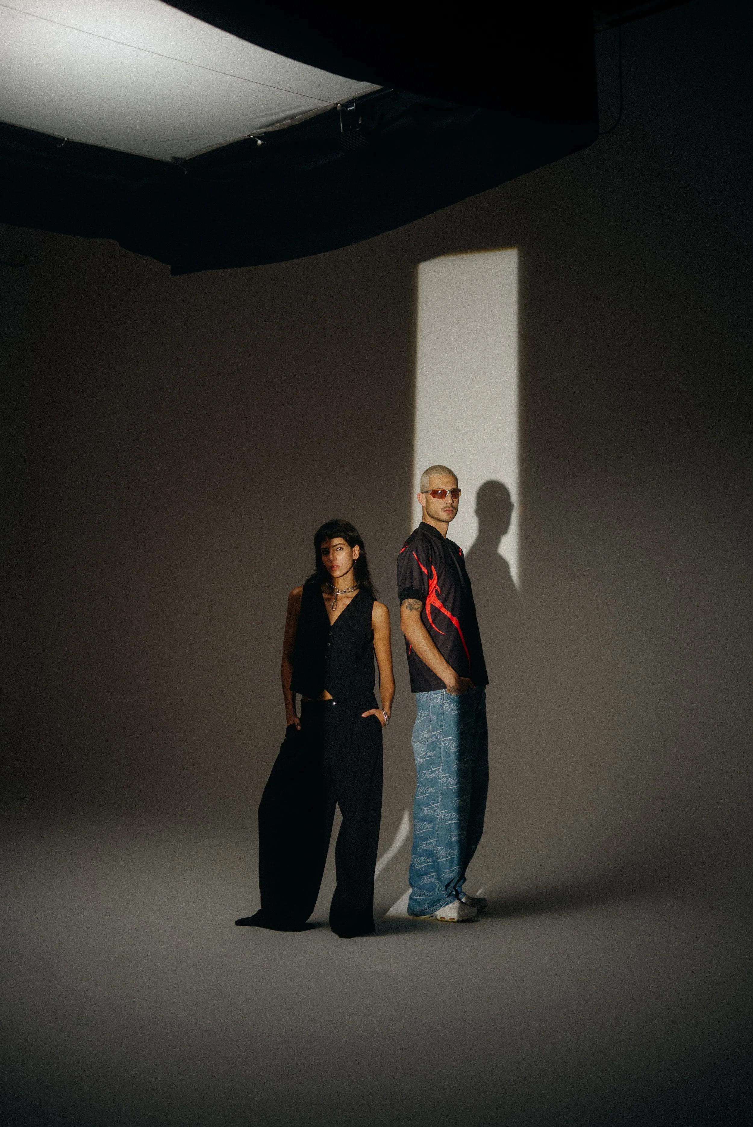 Ein Mann und eine Frau stehen vor einer dunklen neutralen Hintergrund, beleuchtet von einem schrägen Lichtstrahl.