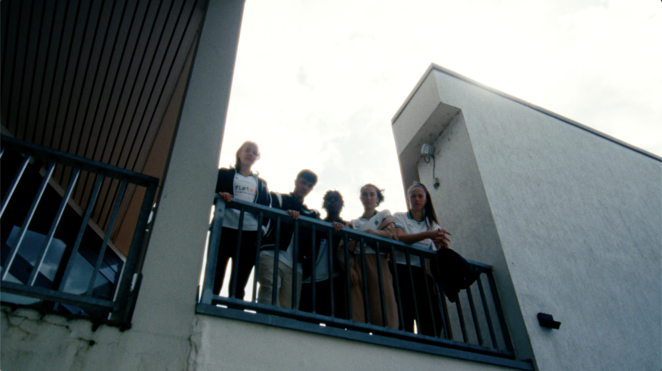 Fünf Jugendliche stehen auf einer Balkonbrüstung und schauen nach unten.
