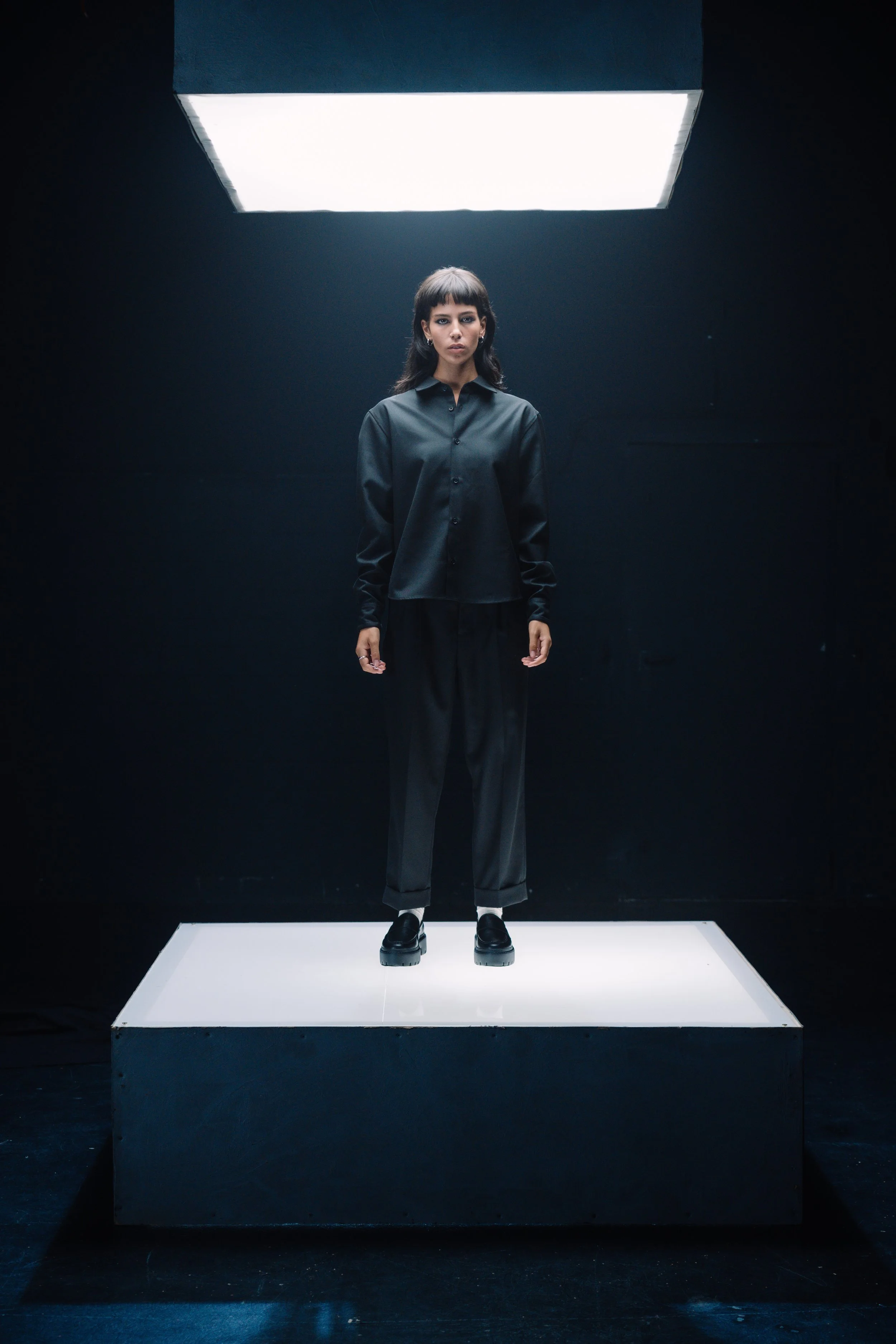 Junge Frau steht auf einem beleuchteten Podest in einem dunklen Raum. Sie trägt eine schwarze Bluse, schwarze Hosen und schwarze Schuhe.