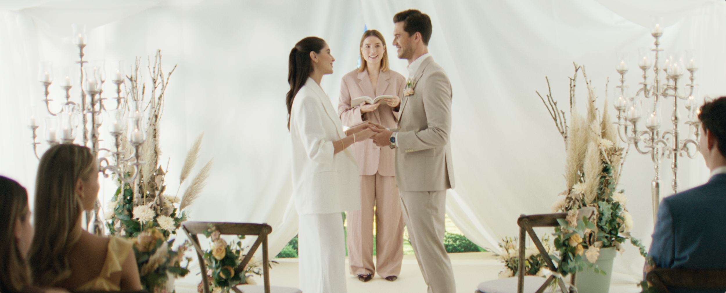 Ein heiratendes Paar tauscht die Ehegelübde während einer Hochzeitszeremonie in einem hell dekorierten Raum mit weißen Vorhängen, Blumen und Kerzen, während die Trauzeugen und Gäste zuschauen.