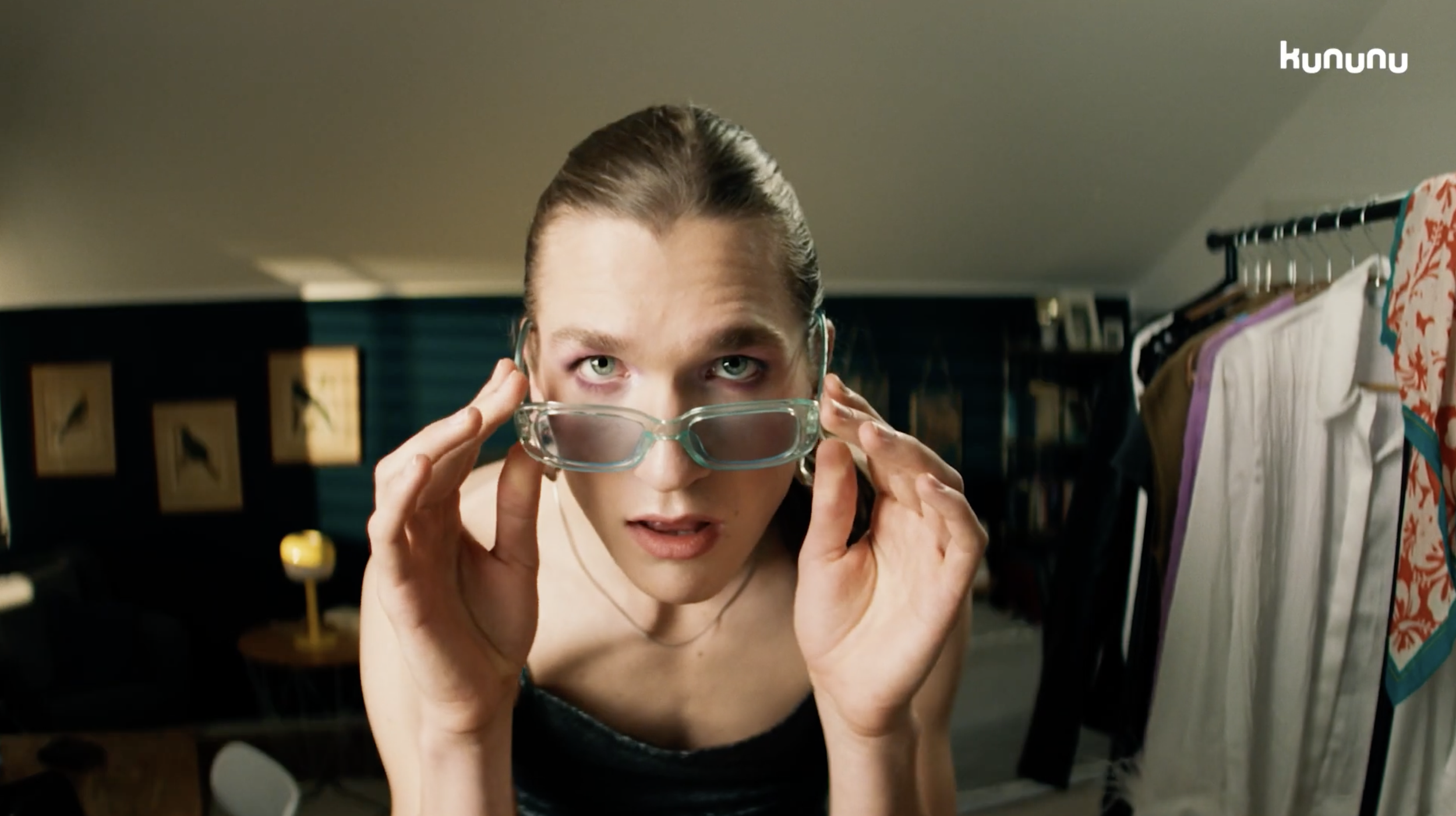 Frau mit Brille, die in einem Raum mit Kleiderstange und Bildern im Hintergrund steht.