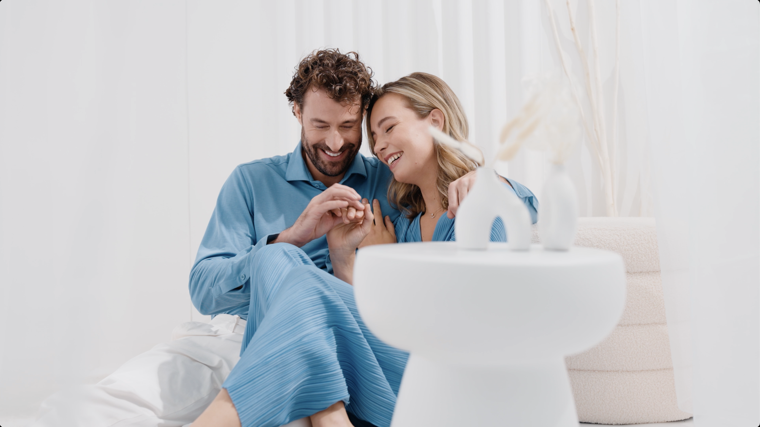 Ein glückliches Paar in blauen Kleidern, das auf einem Bett sitzt, hält sich an den Händen und lächelt.