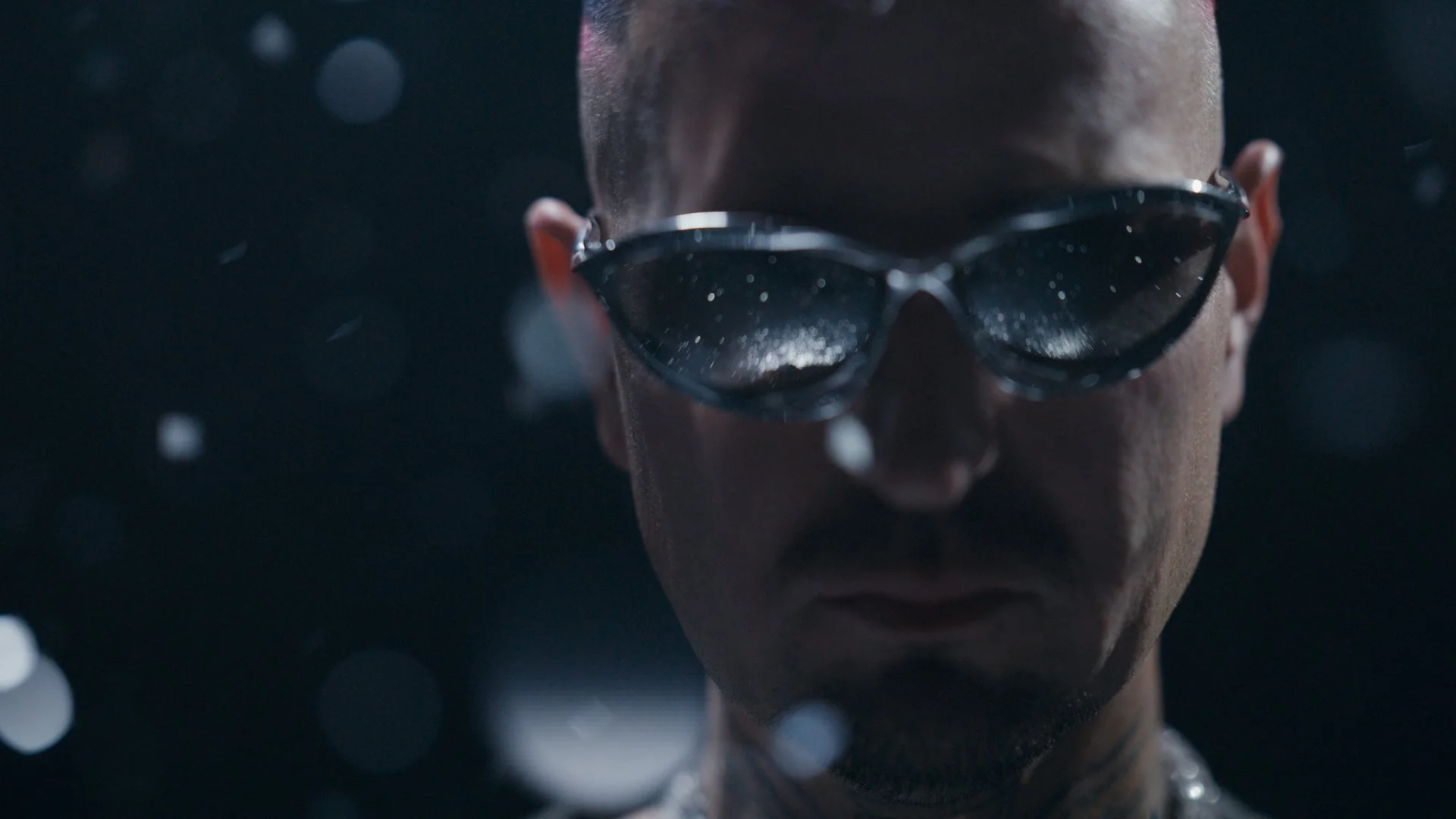 Nahaufnahme von Robin Schulz, der dunkle Sonnenbrille trägt und in einem Wasser- oder Schneegebiet steht, mit Wassertröpfchen auf seiner Haut und Oberflächenreflexionen in den Gläsern der Brille.