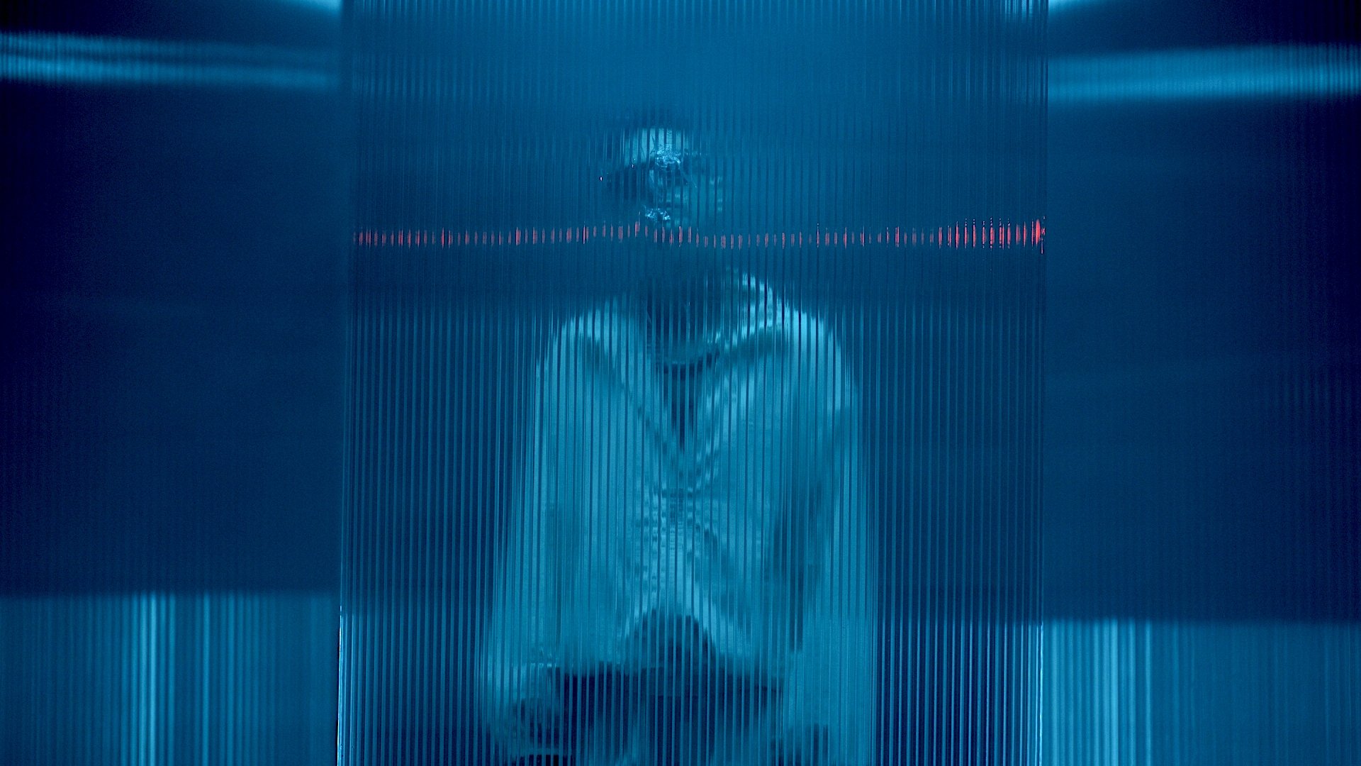 Robin Schulz, der in einem Raum mit blauer Beleuchtung hinter einer durchsichtigen, geriffelten Glaswand steht.