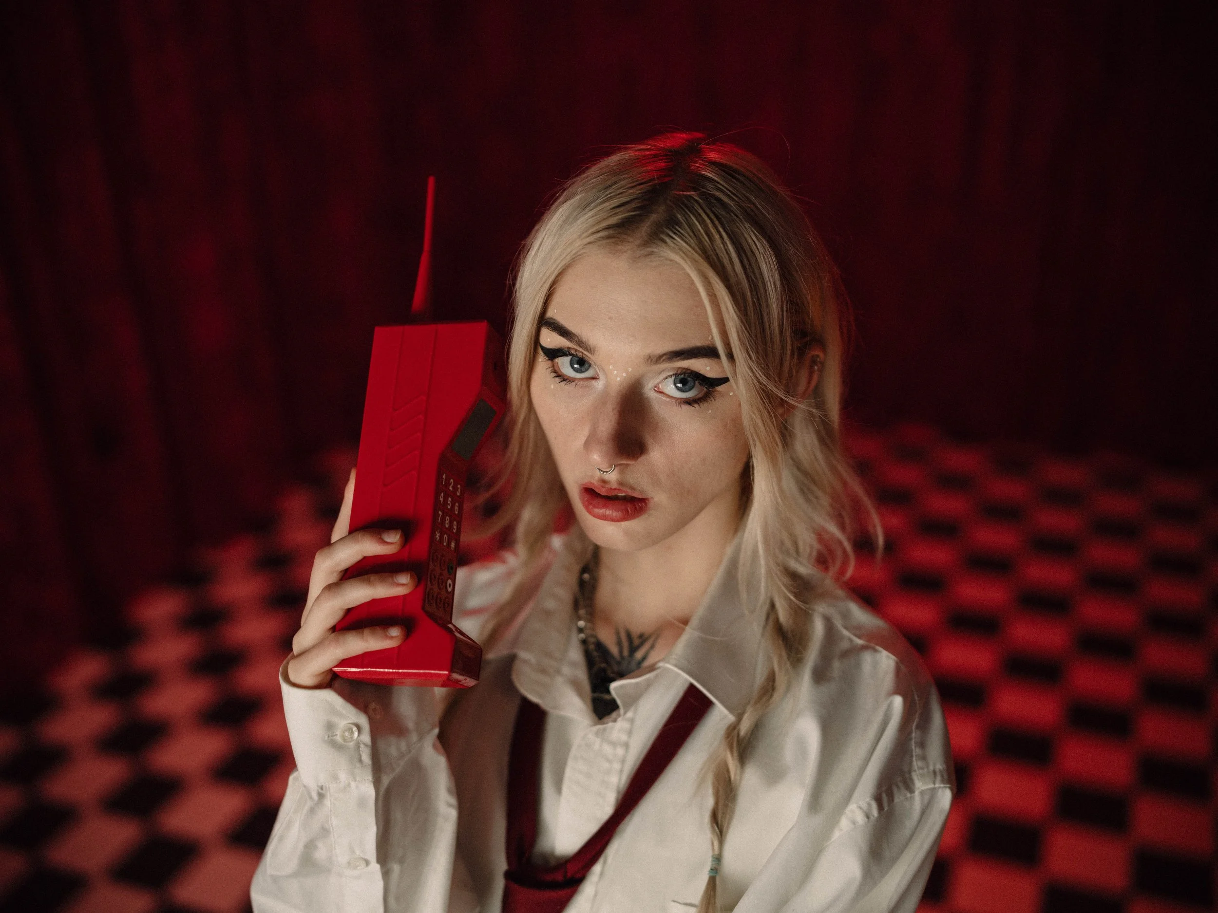 Junge Frau mit blondem Haar, blauen Augen, rotem Lippenstift, trägt eine weiße Bluse mit Krawatte, hält einen roten alten Telefont in der Hand, im Hintergrund dunkler Raum mit rotem und schwarzem Fliesenboden.