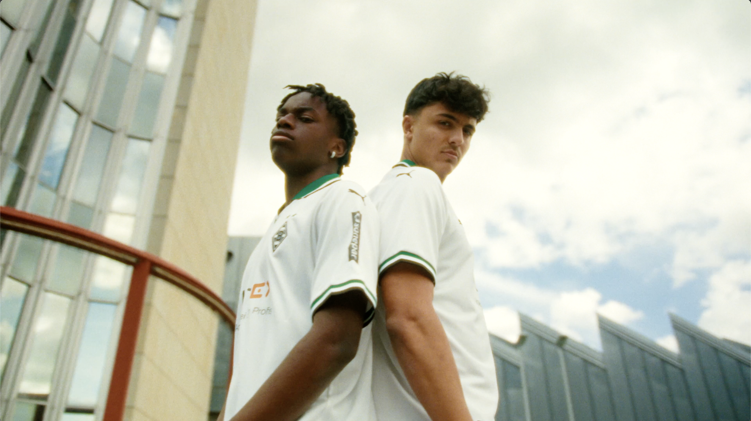 Zwei junge Männer stehen Rücken an Rücken vor moderner Architektur mit Glas und Metall, tragen weiße Trikots und schauen ernst in die Kamera bei Sonnenwetter, blauer Himmel mit Wolken im Hintergrund.