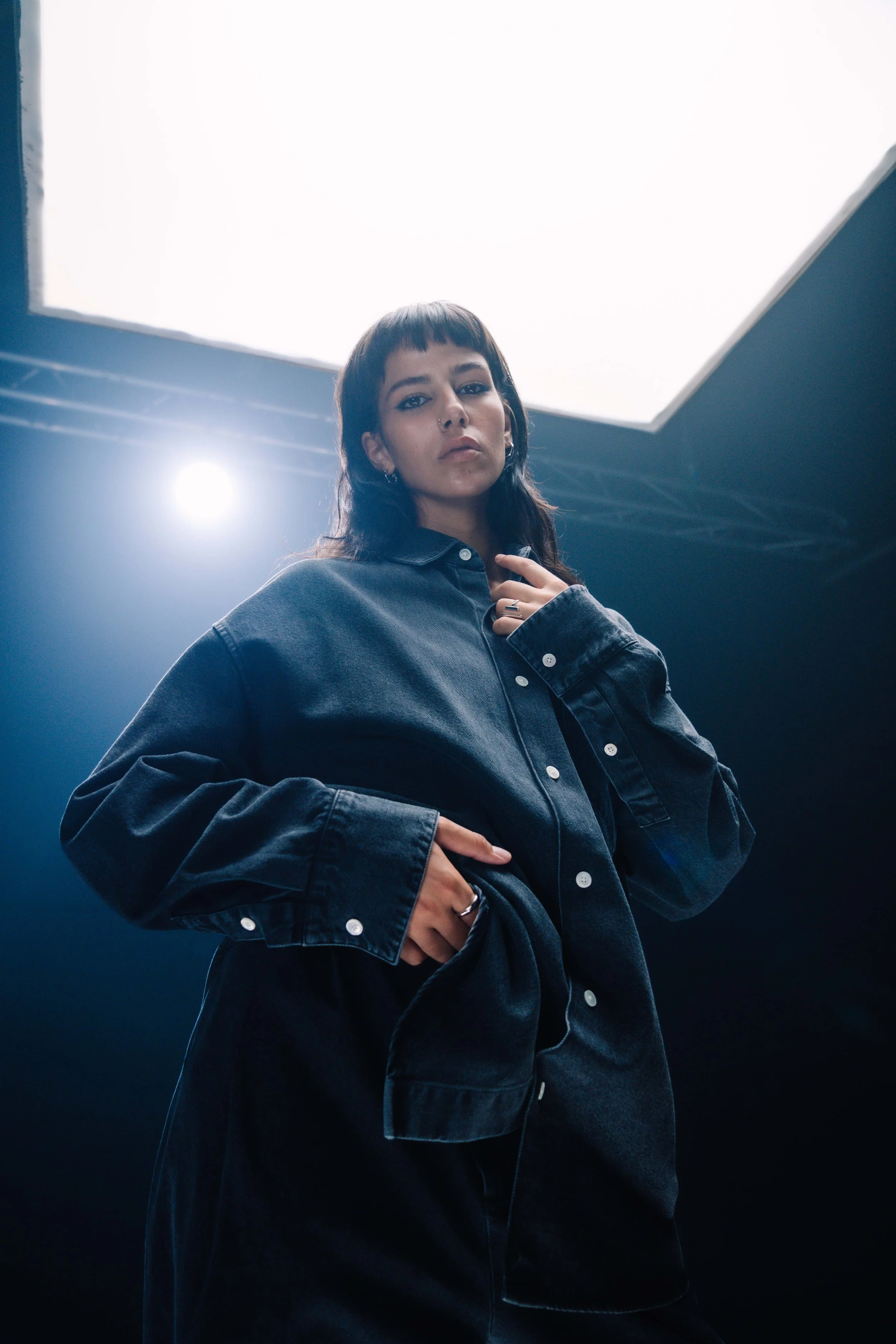 Junge Frau in dunkler Denim-Jacke vor hell beleuchtetem Hintergrund, blickt ernst in die Kamera.