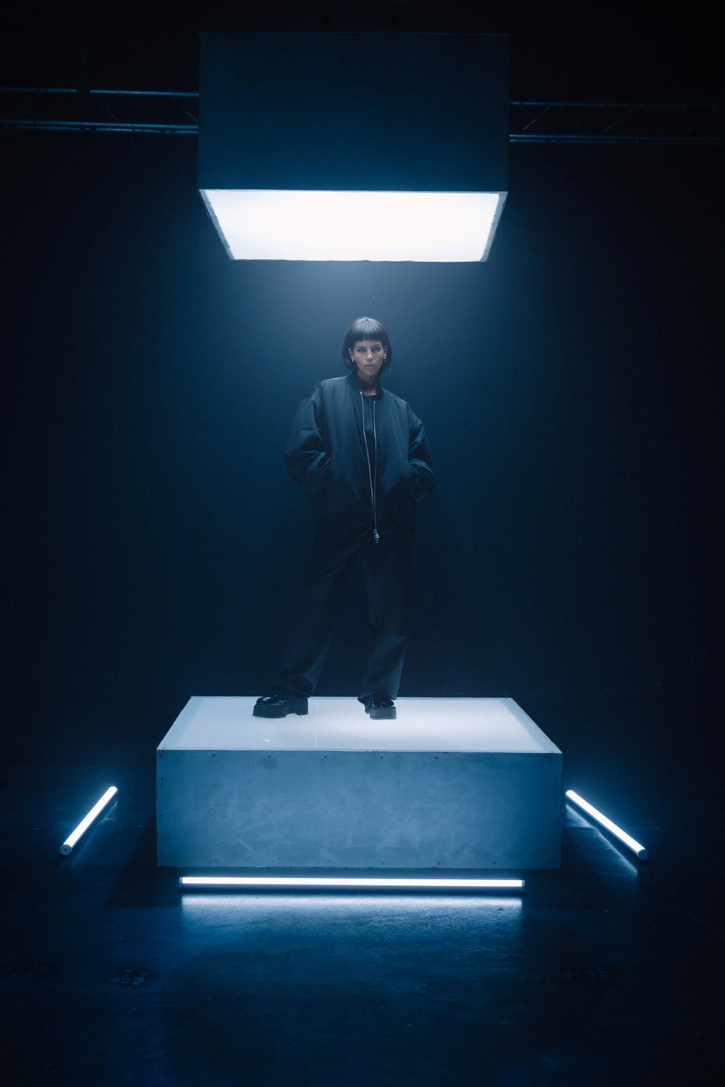 Junge Frau steht auf einer weißen Plattform unter einer großen, leuchtenden Lichteinheit, in einer dunklen Umgebung mit bläulichem Licht, trägt dunkle Kleidung und schaut direkt in die Kamera.