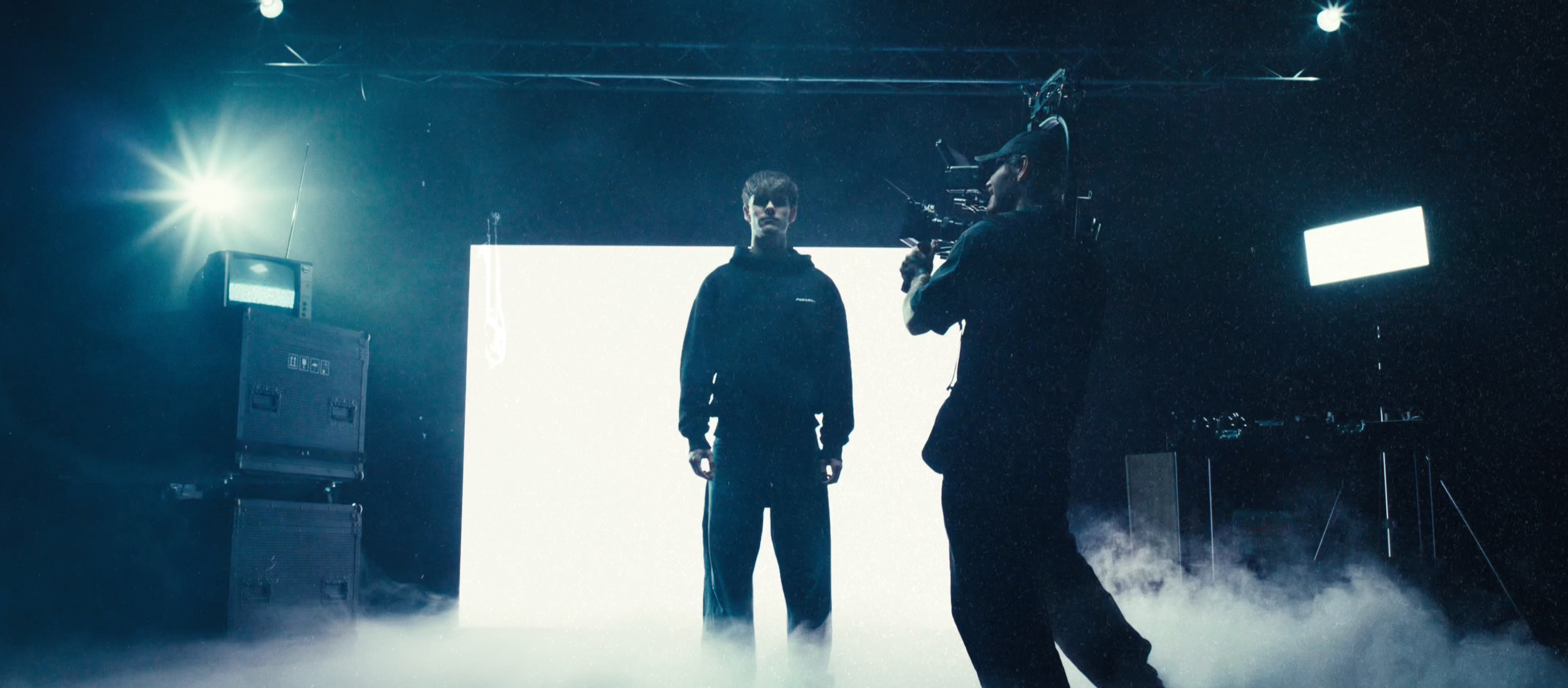 Ein Mann steht vor einem weißen Hintergrund auf einer Bühne, während ein Videograf ihn filmt, im dunklen Raum mit Scheinwerfern und Nebel.