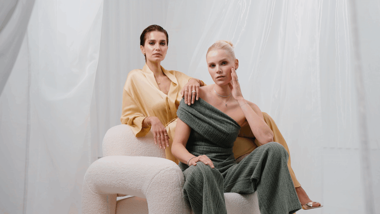 Zwei Frauen sitzen auf einem modernen Stuhl vor weißen Vorhängen, eine in einem beigen Satin-Look und die andere in einem grauen, off-shoulder Kleid, beide wirken nachdenklich.