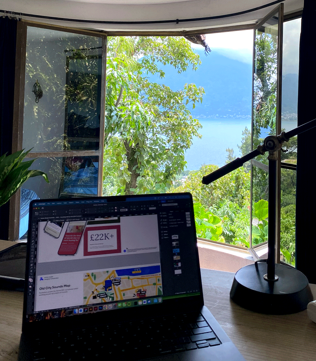 Open window overlooking lush greenery and a large body of water with mountains in the distance, viewed from a desk with a laptop and a black desk lamp.
