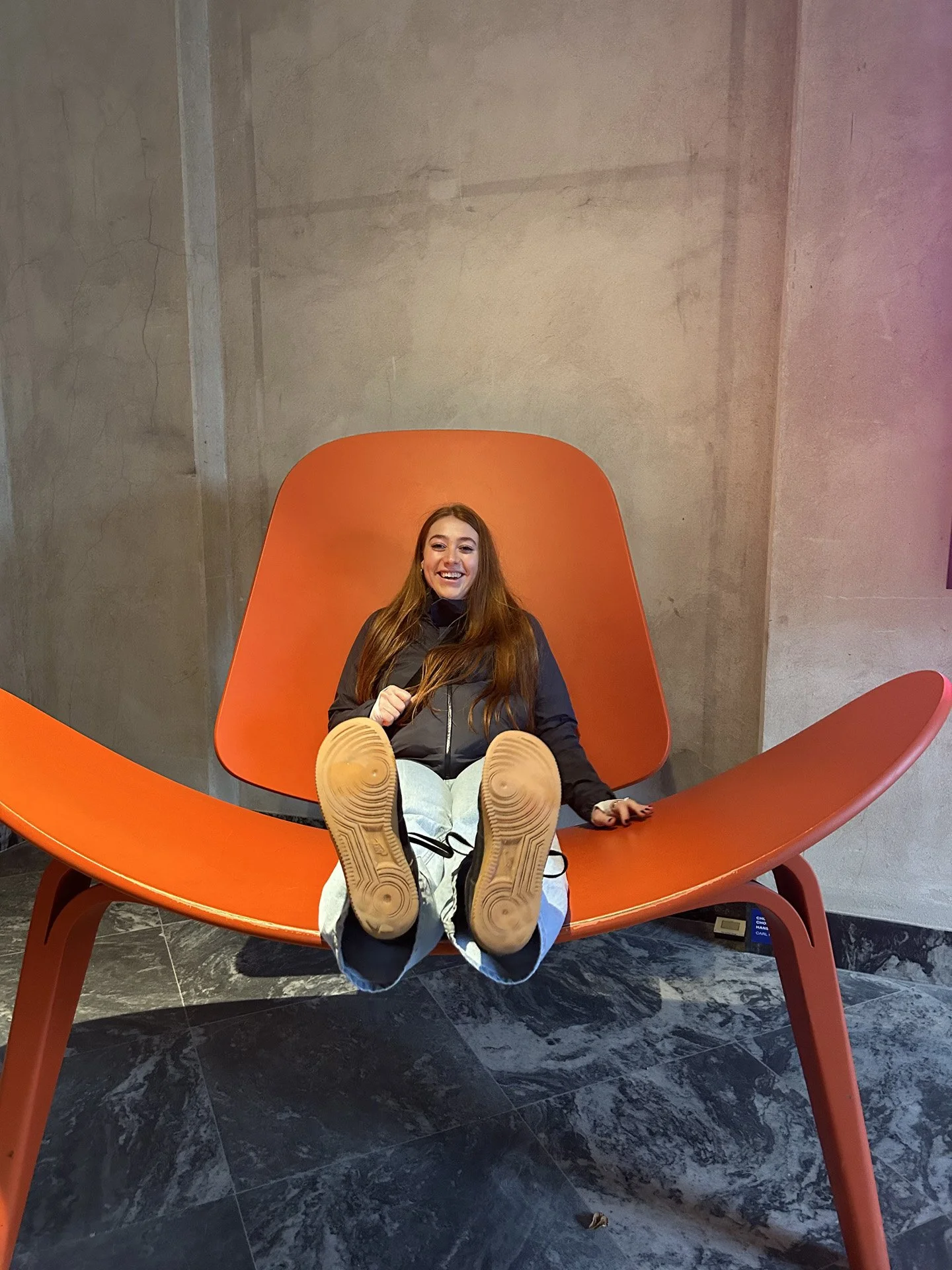 A young woman with long red hair, smiling and sitting on an oversized, orange chair, in an art gallery exhibit with concrete walls and a marble floor.