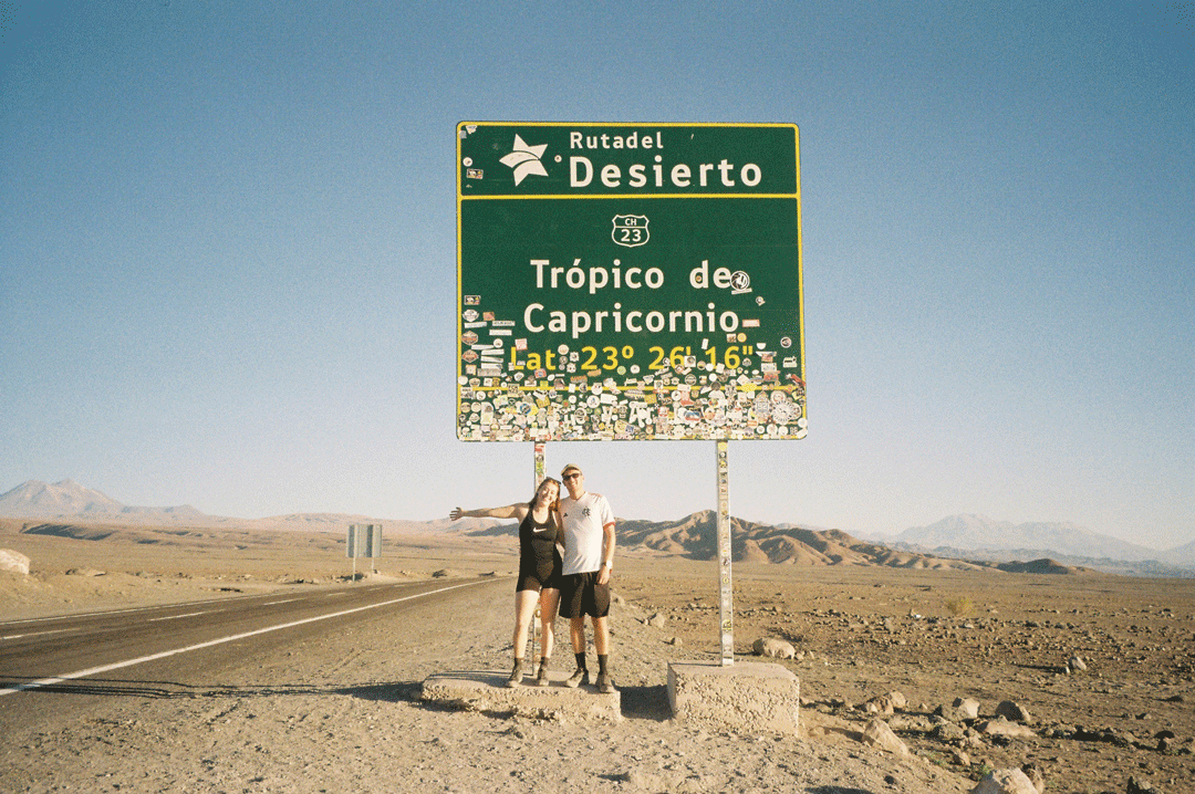 A woman and man standing next to a large green road sign in a desert landscape, with distant mountains and a clear blue sky.