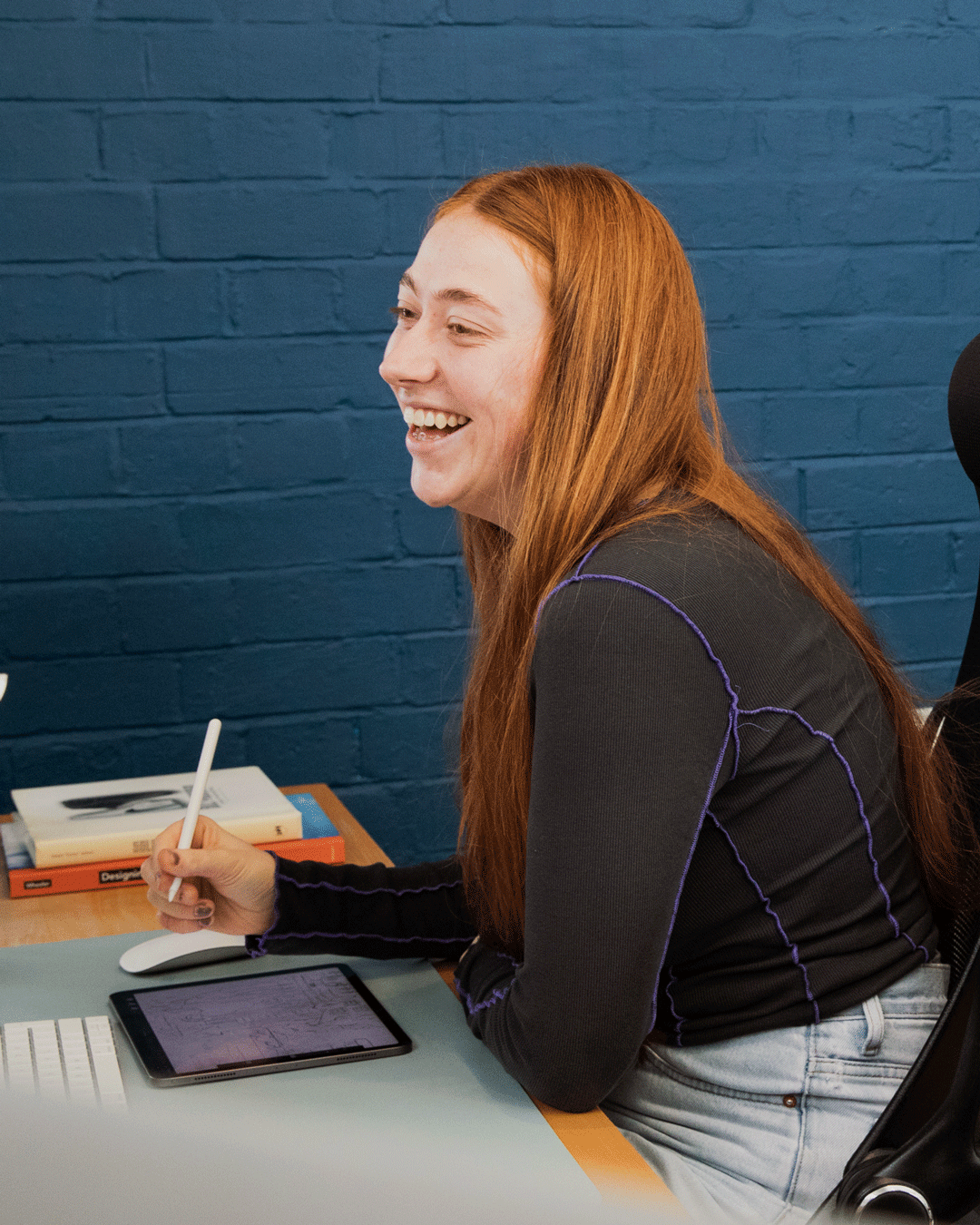 A young woman with red hair smiling and working at a desk with a tablet, keyboard, and a computer mouse in front of a blue brick wall.