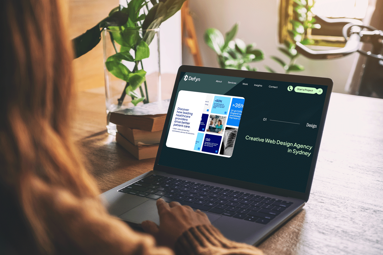 A woman working on a laptop at a wooden desk, with a potted plant and books visible in the background, displaying a web design agency's website.