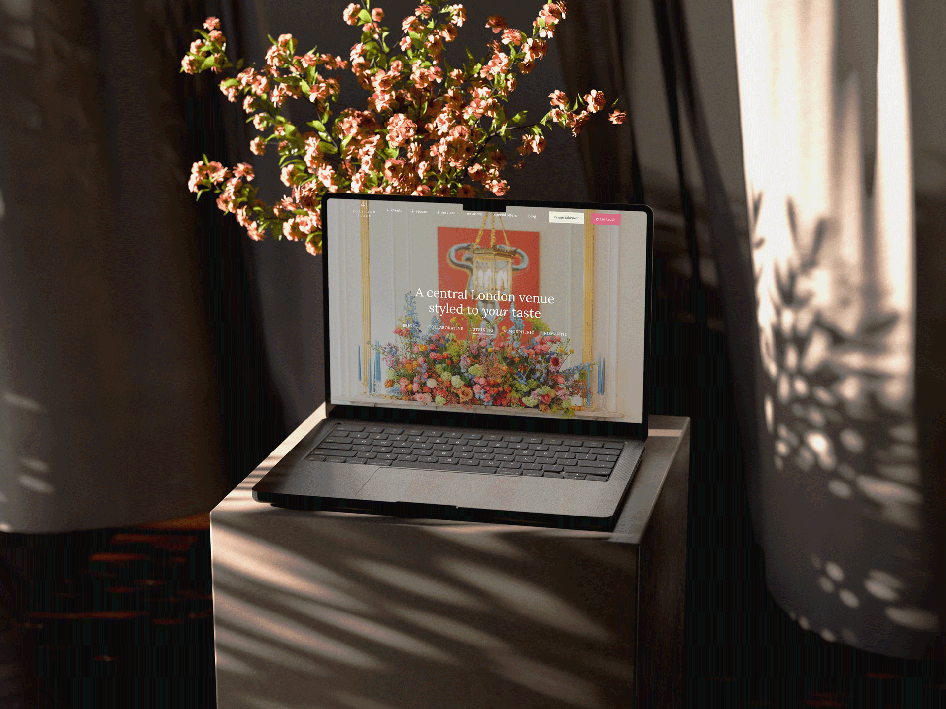 A laptop displaying a website with a colorful floral arrangement in the background, placed on a wooden table with sunlight filtering through curtains creating shadows.
