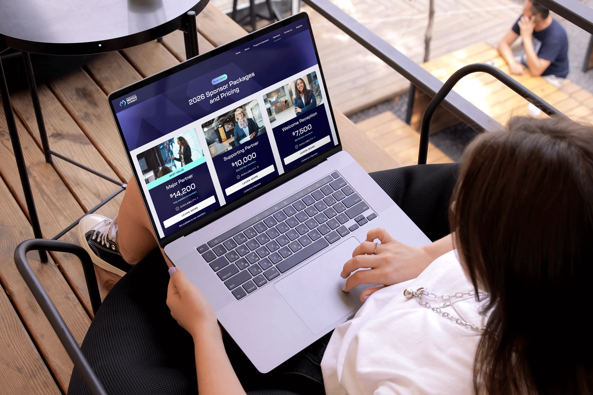 A person sitting on a bench using a MacBook laptop at an outdoor wooden patio, viewing a webpage about sponsor packages and pricing.