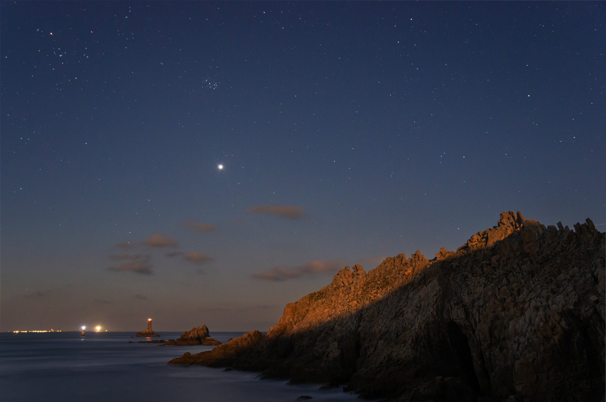 Nighttime coastal scene with rocky cliffs, a lighthouse with a bright light, and a starry sky with several bright planets or stars.