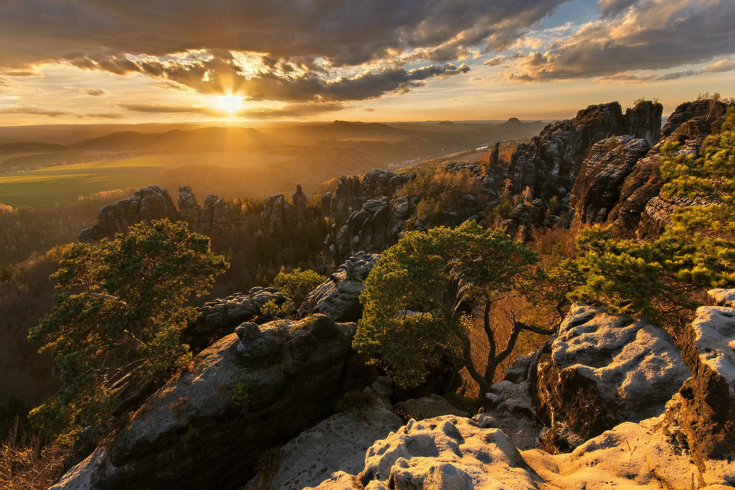 Sunset over rocky cliffs and sparse trees in a mountainous landscape, with a valley and fields in the distance. Saxony Switzerland, sächiwsche Schweiz.