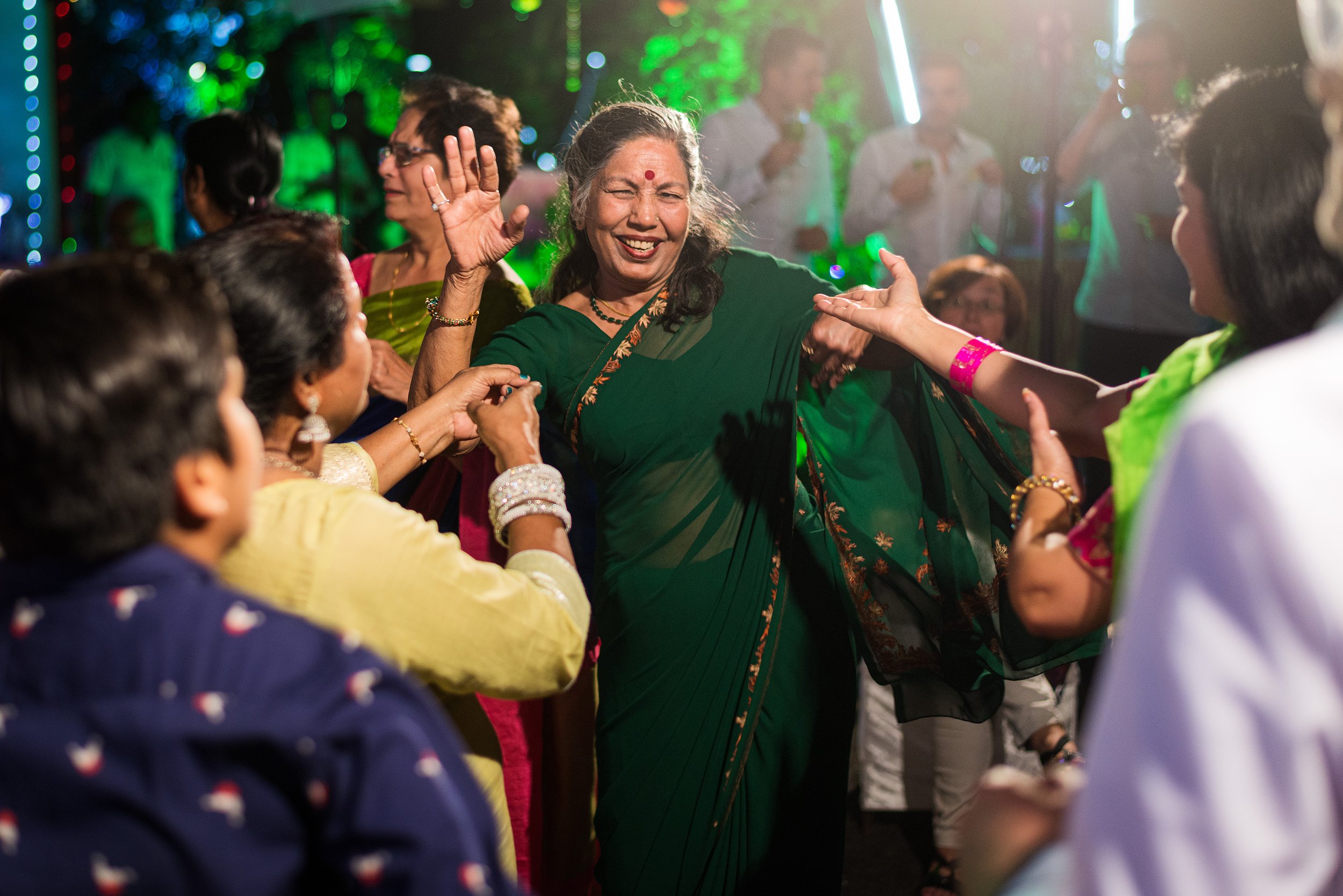 Women dancing and celebrating at an indoor party with colorful lighting, wearing traditional Indian clothing.